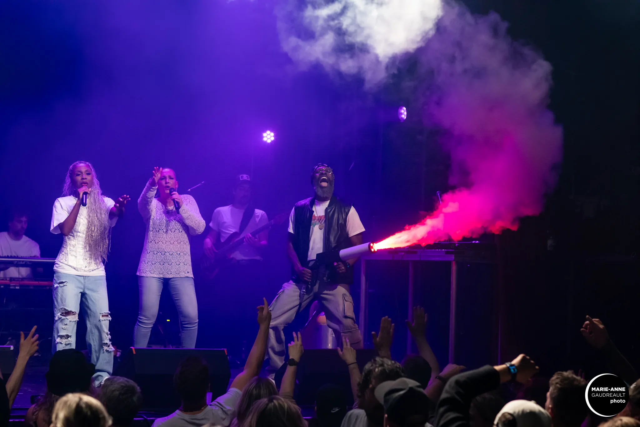 Live concert with two female singers and a male performer shooting colorful smoke into the crowd amid purple stage lighting.