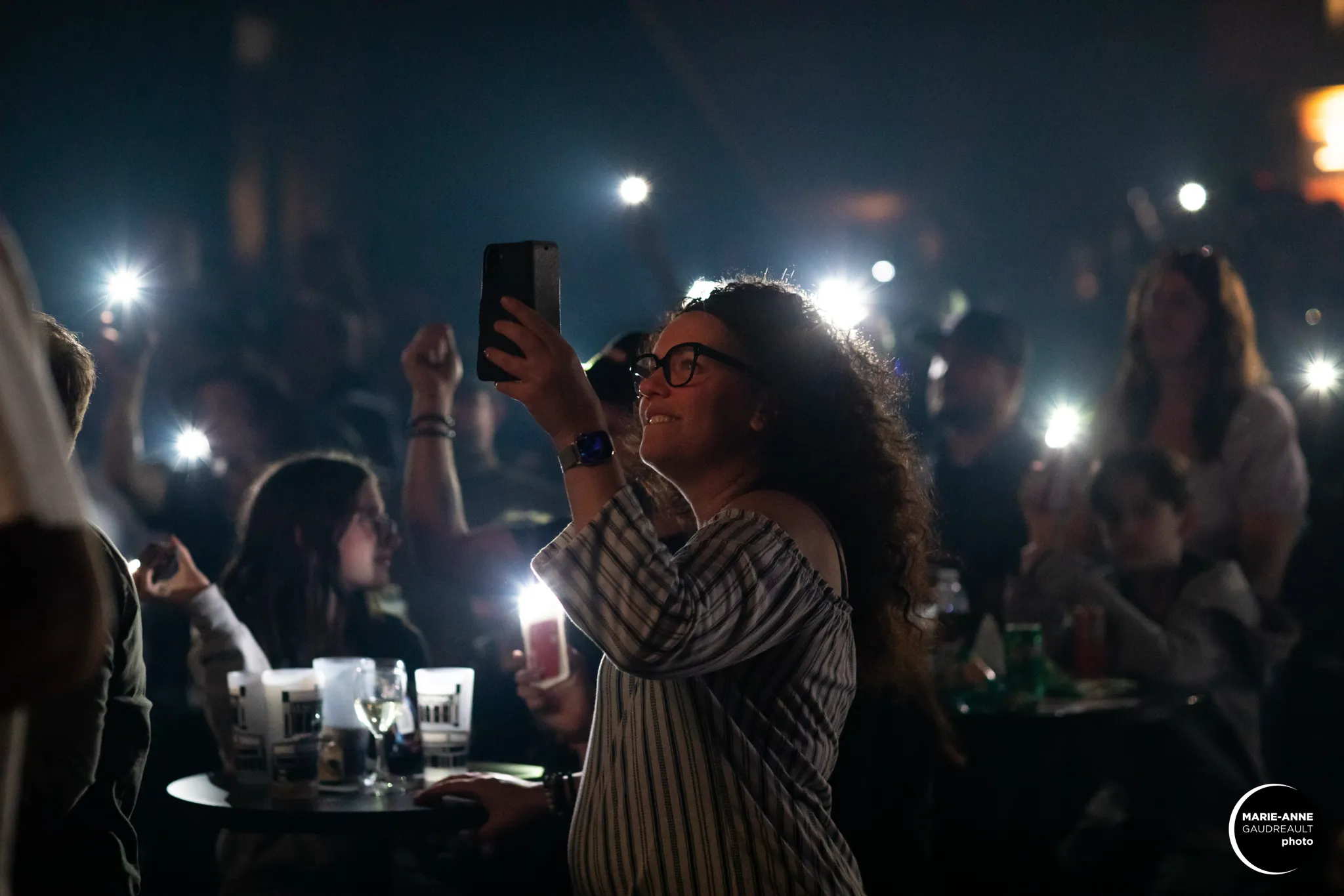 Woman with curly hair and glasses holding up a smartphone in a dimly lit crowd with many people using phone flashlights.