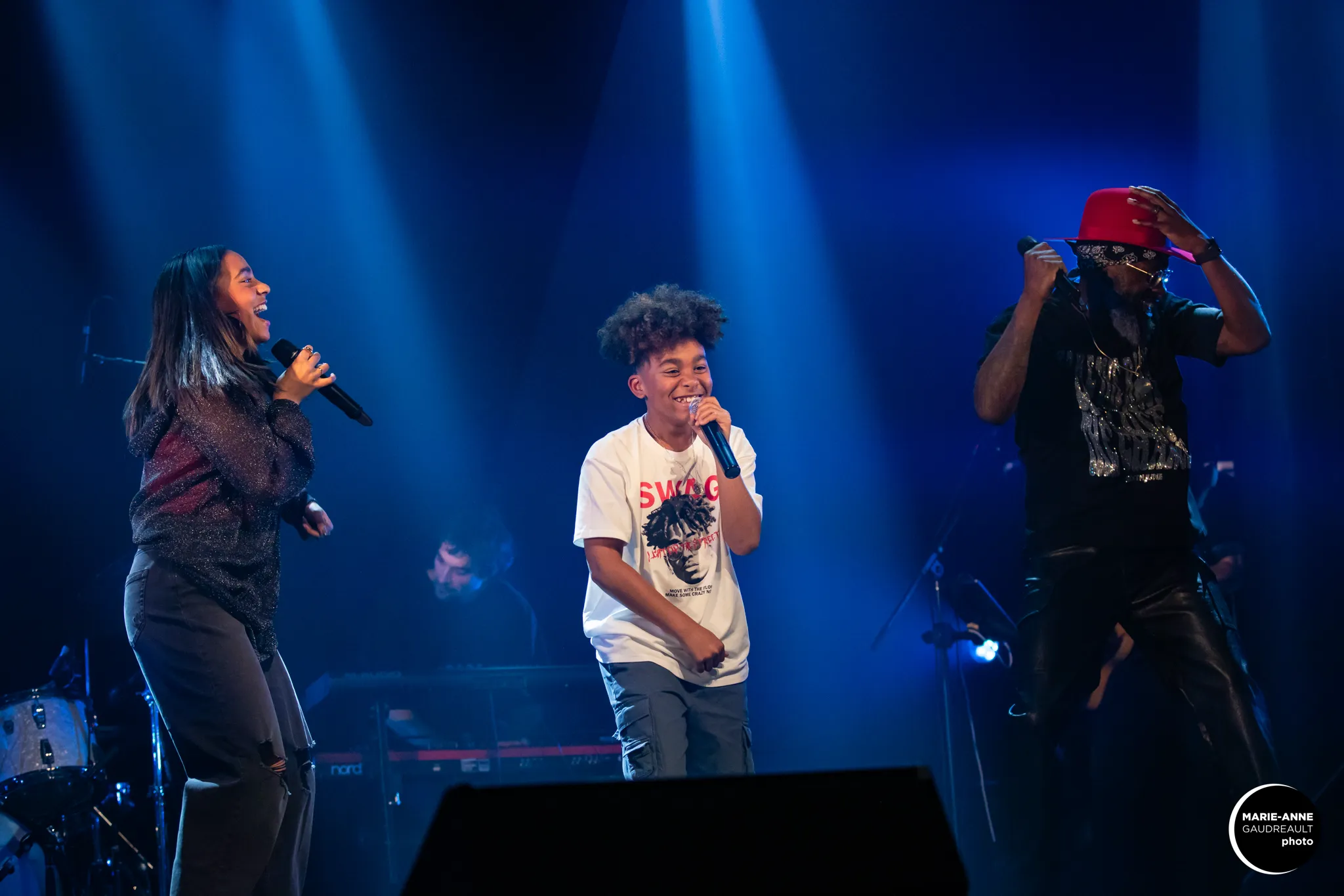 Three performers singing and dancing on stage under blue spotlights, with a keyboard player in the background.