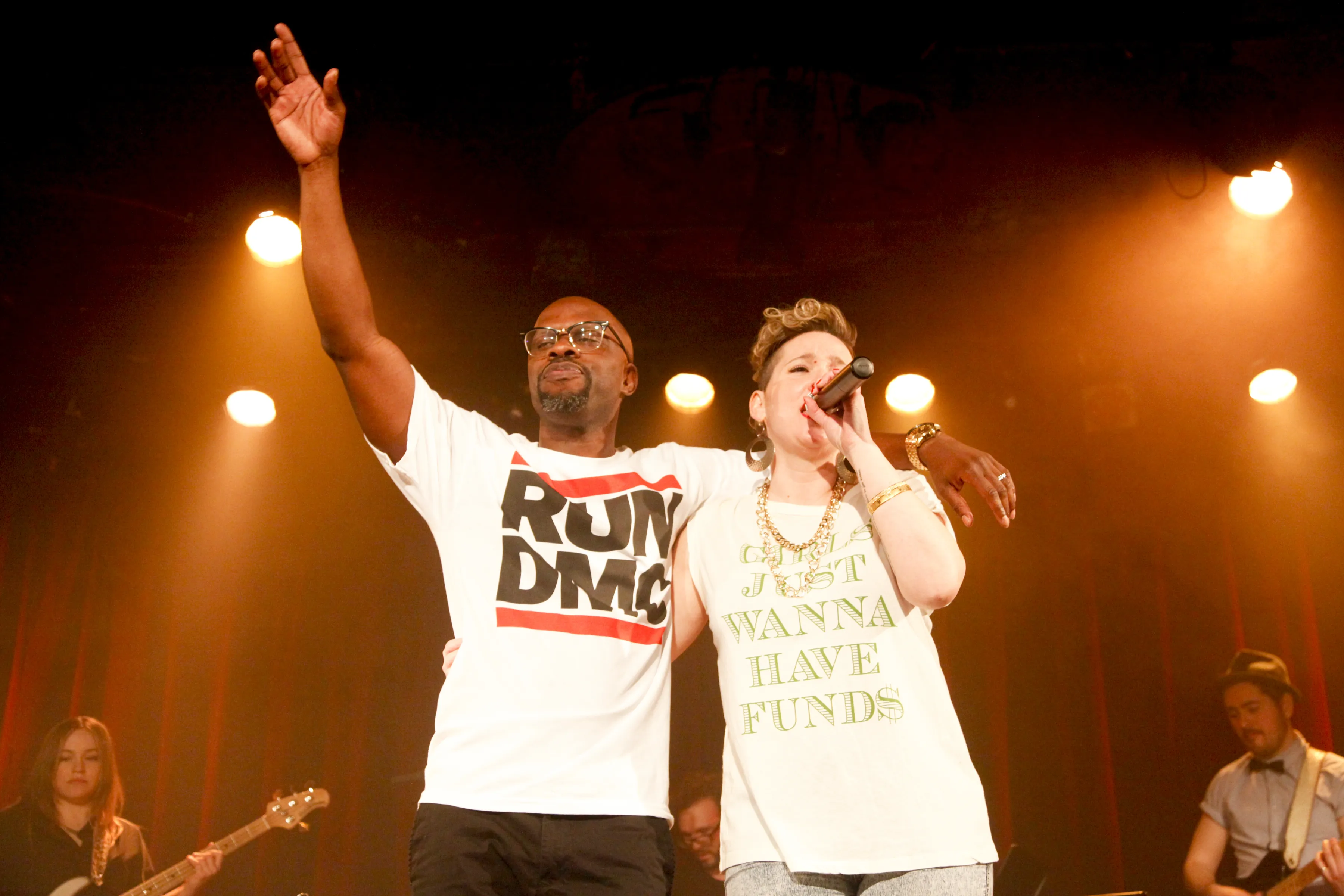 Two performers on stage, one wearing a Run DMC t-shirt raising his arm, and the other singing into a microphone wearing a shirt that says 'Just Wanna Have Funds'.