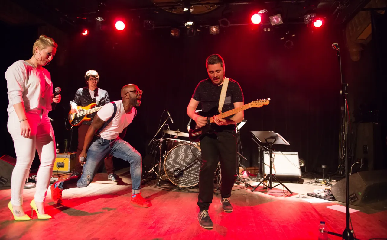 Band performing on stage with guitarist, bassist, singer in white outfit and energetic vocalist in red shoes under red stage lights.