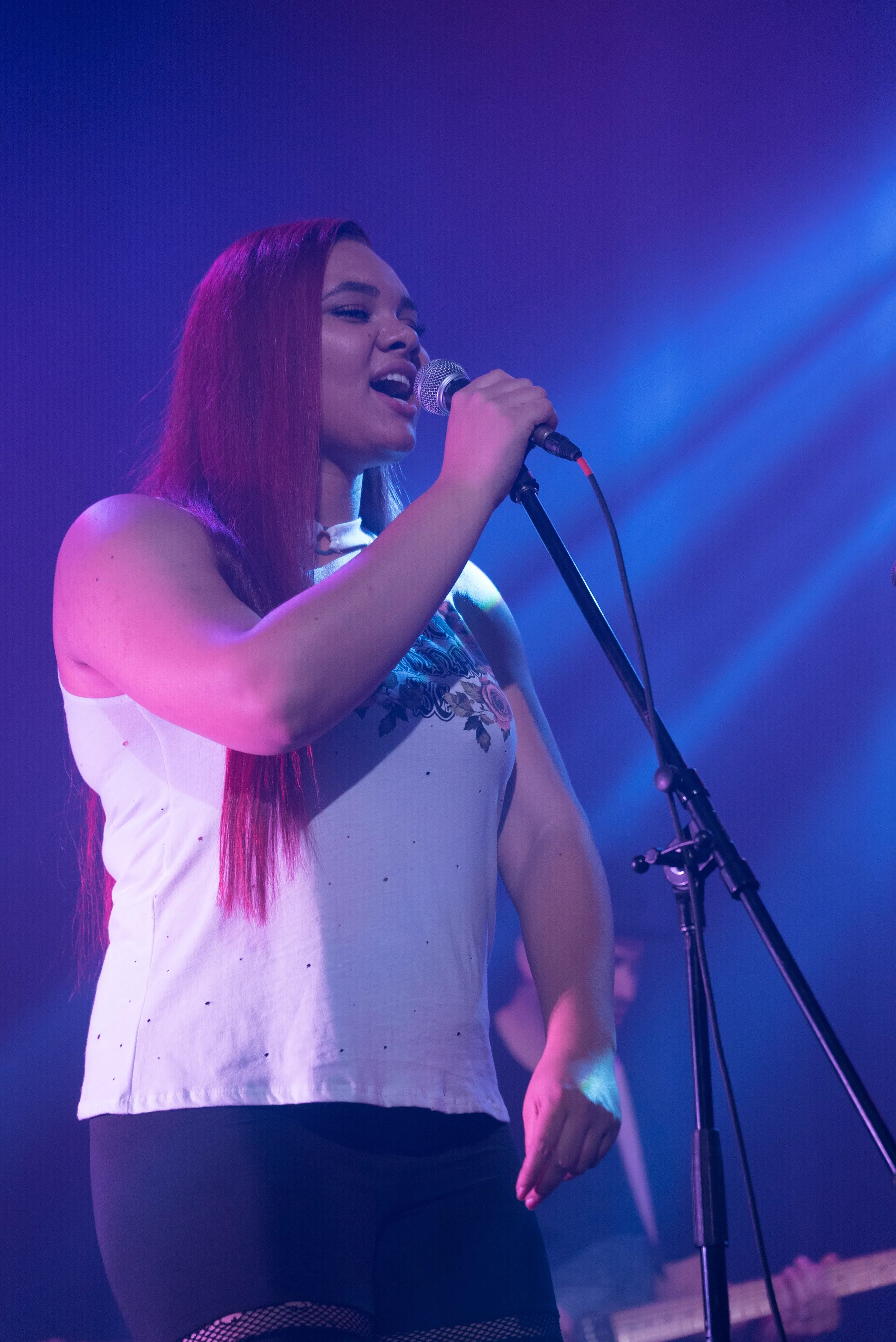 Woman with long red hair singing into a microphone on stage under purple and blue lights.