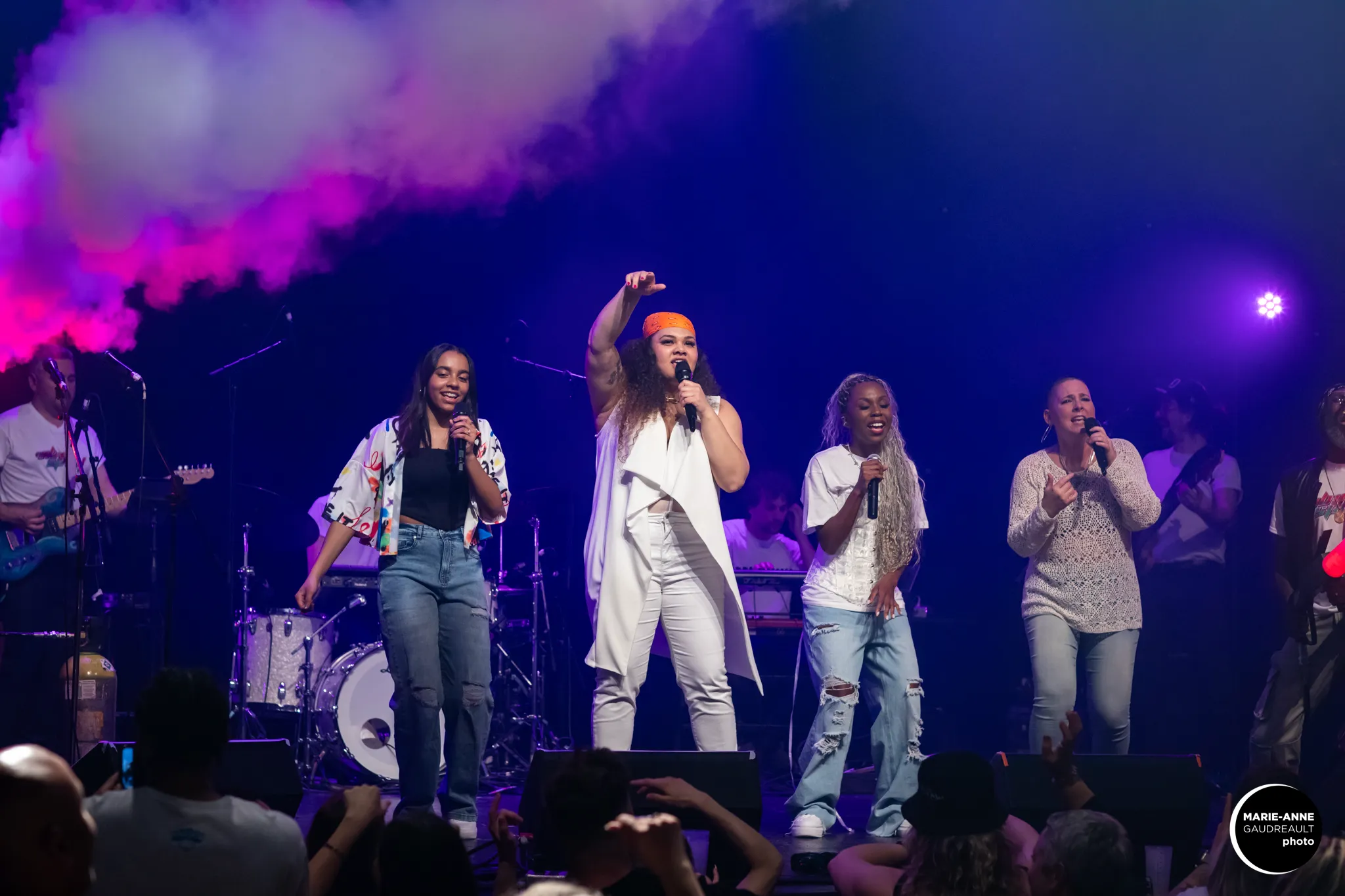 Four singers performing on stage with microphones amid purple and pink smoke effects, accompanied by musicians in the background.