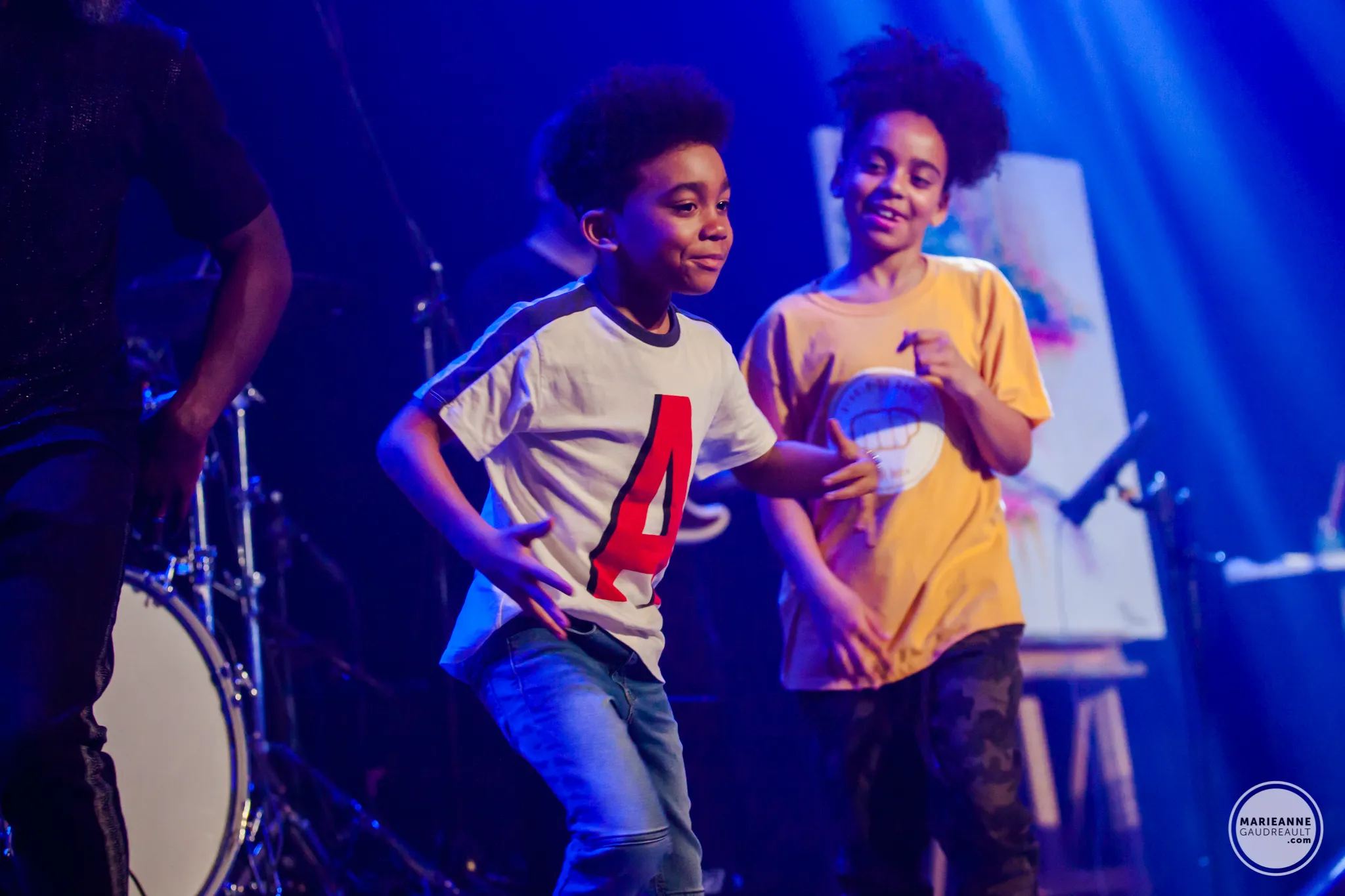 Two children dancing on a stage under blue lighting with a drum set in the background.