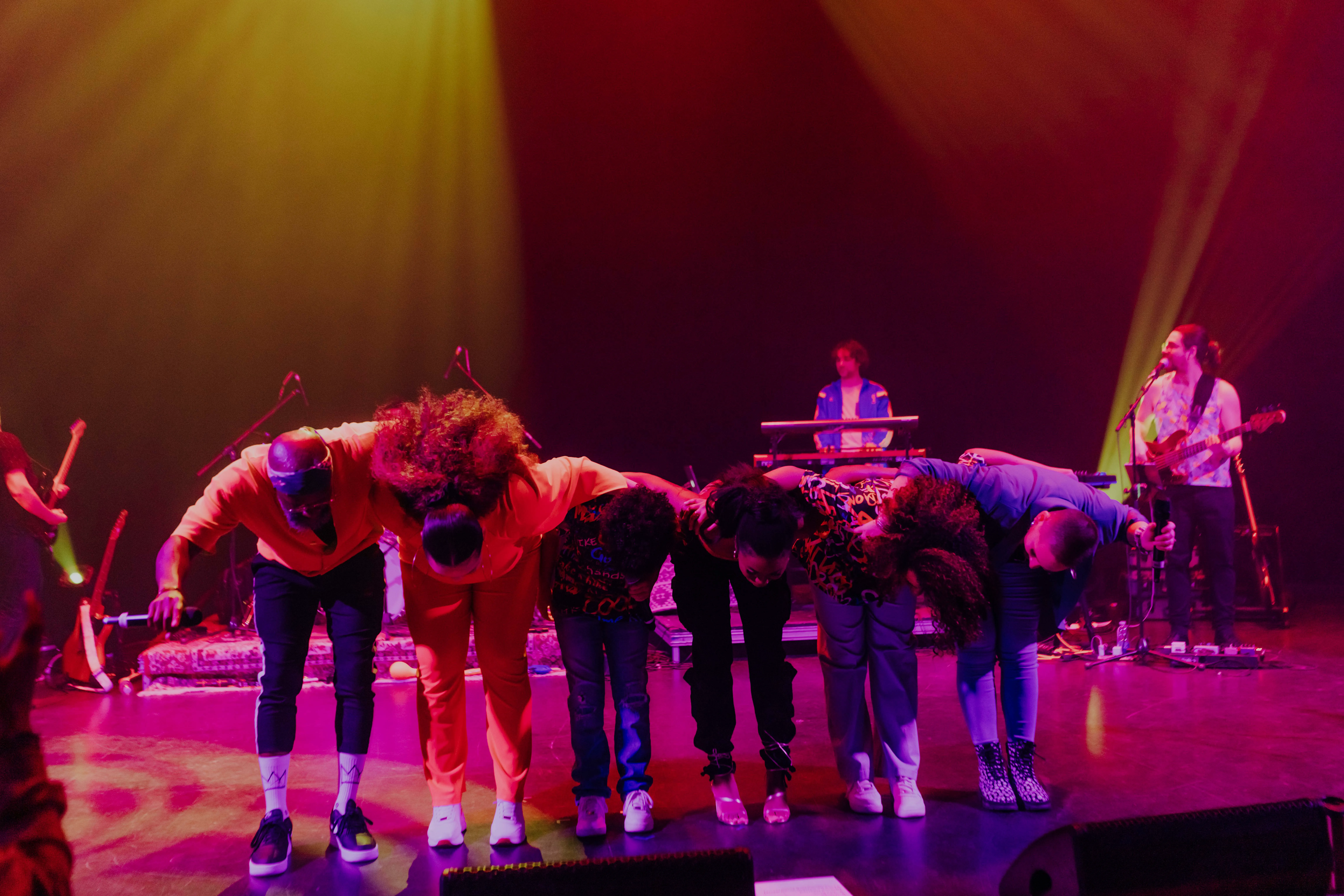 Six performers bowing on stage with colorful lighting, while two musicians play instruments in the background.