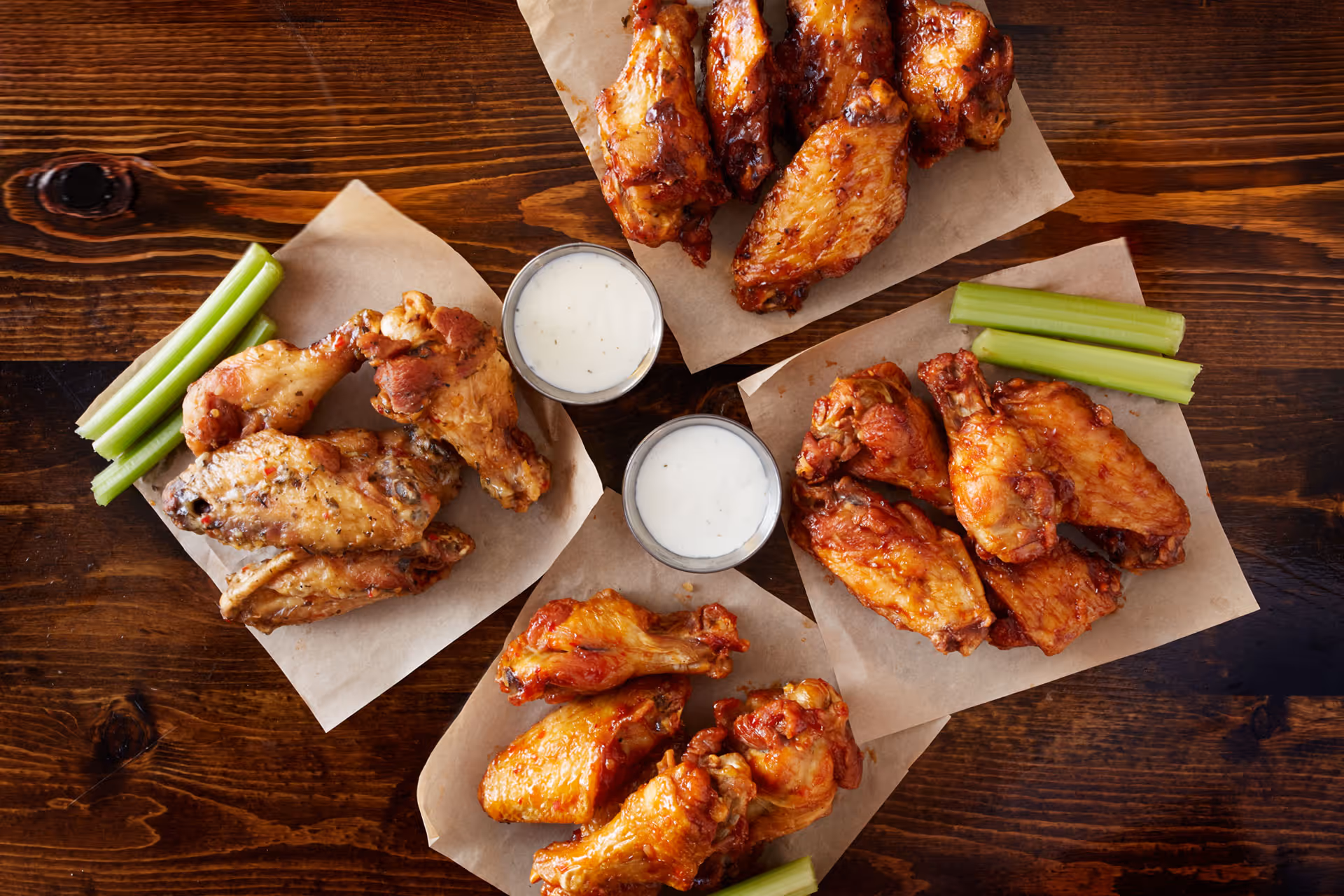 Four servings of chicken wings on parchment paper with celery sticks and two cups of dipping sauce on a wooden table.