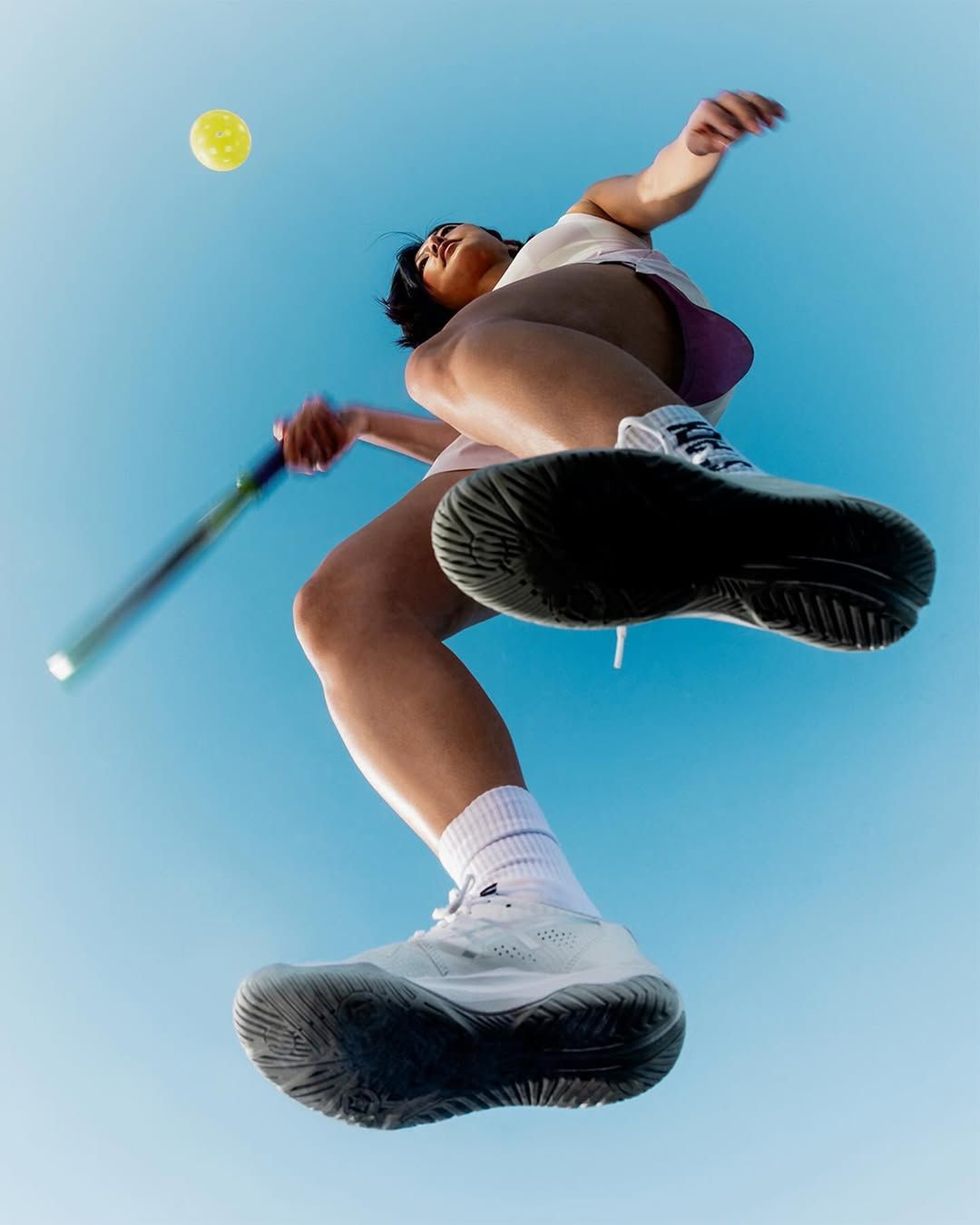 Low-angle view of a woman in athletic wear playing pickleball, swinging a paddle at a yellow ball against a clear blue sky.