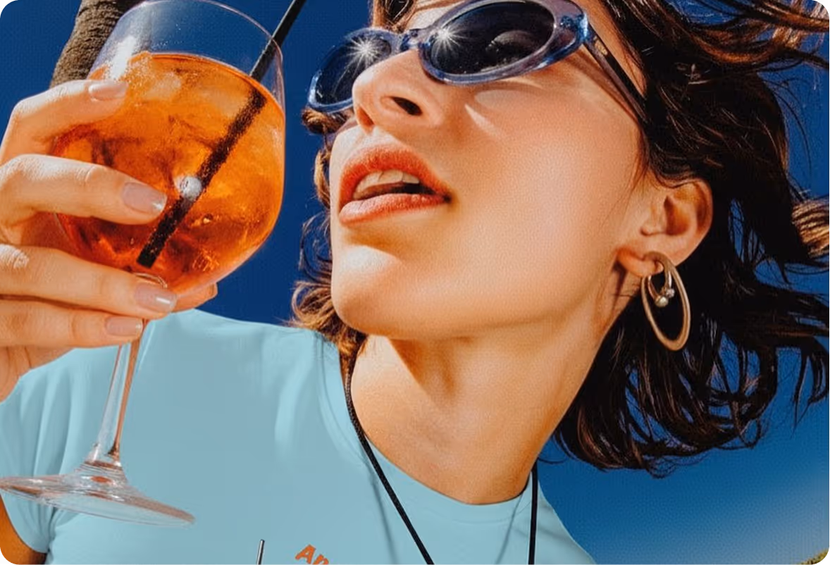Woman wearing sunglasses and hoop earrings holding a glass of orange iced drink against a clear blue sky.