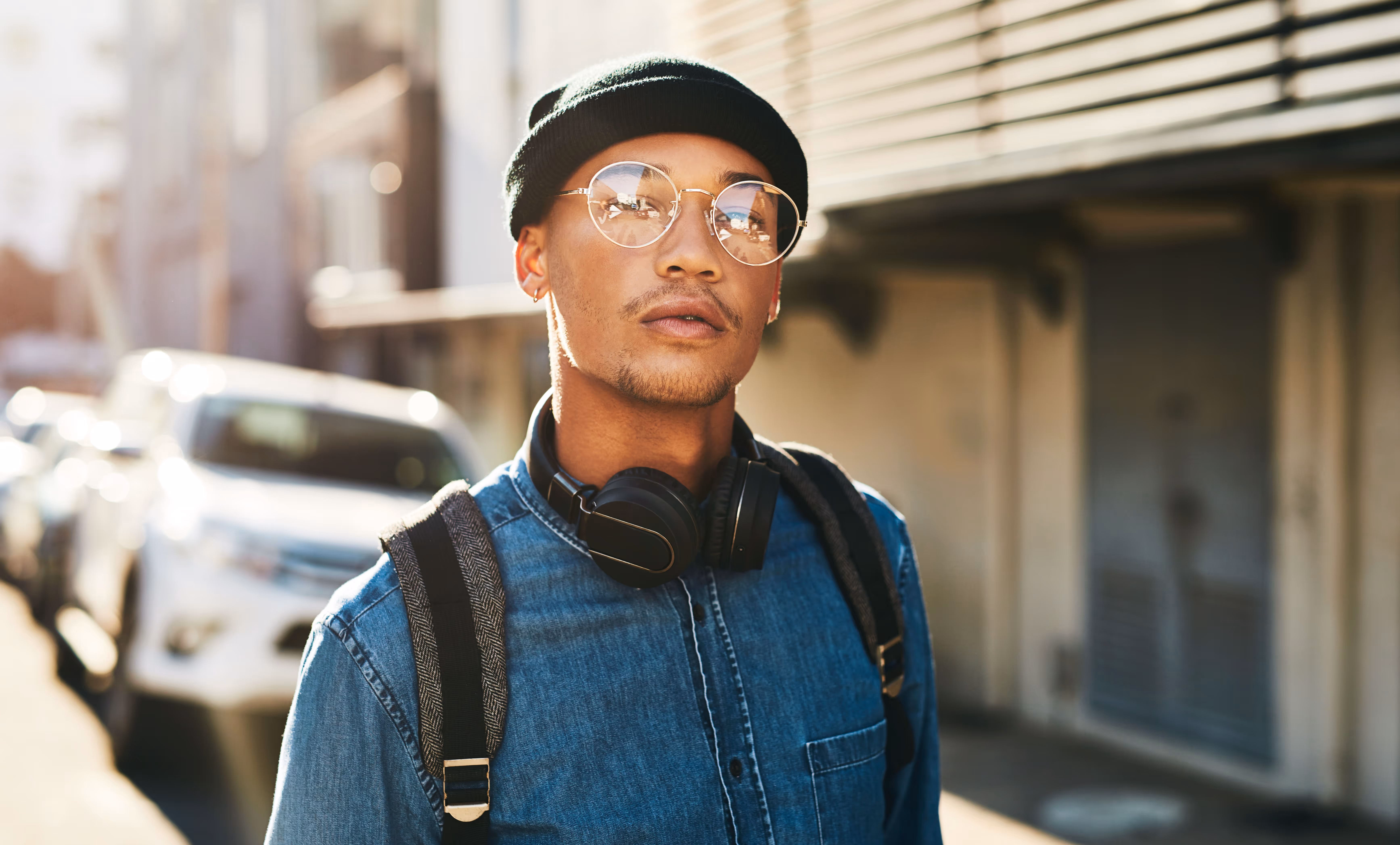 Young man wearing a black beanie, round glasses, headphones around his neck, and a denim shirt standing outdoors with a blurred street background.