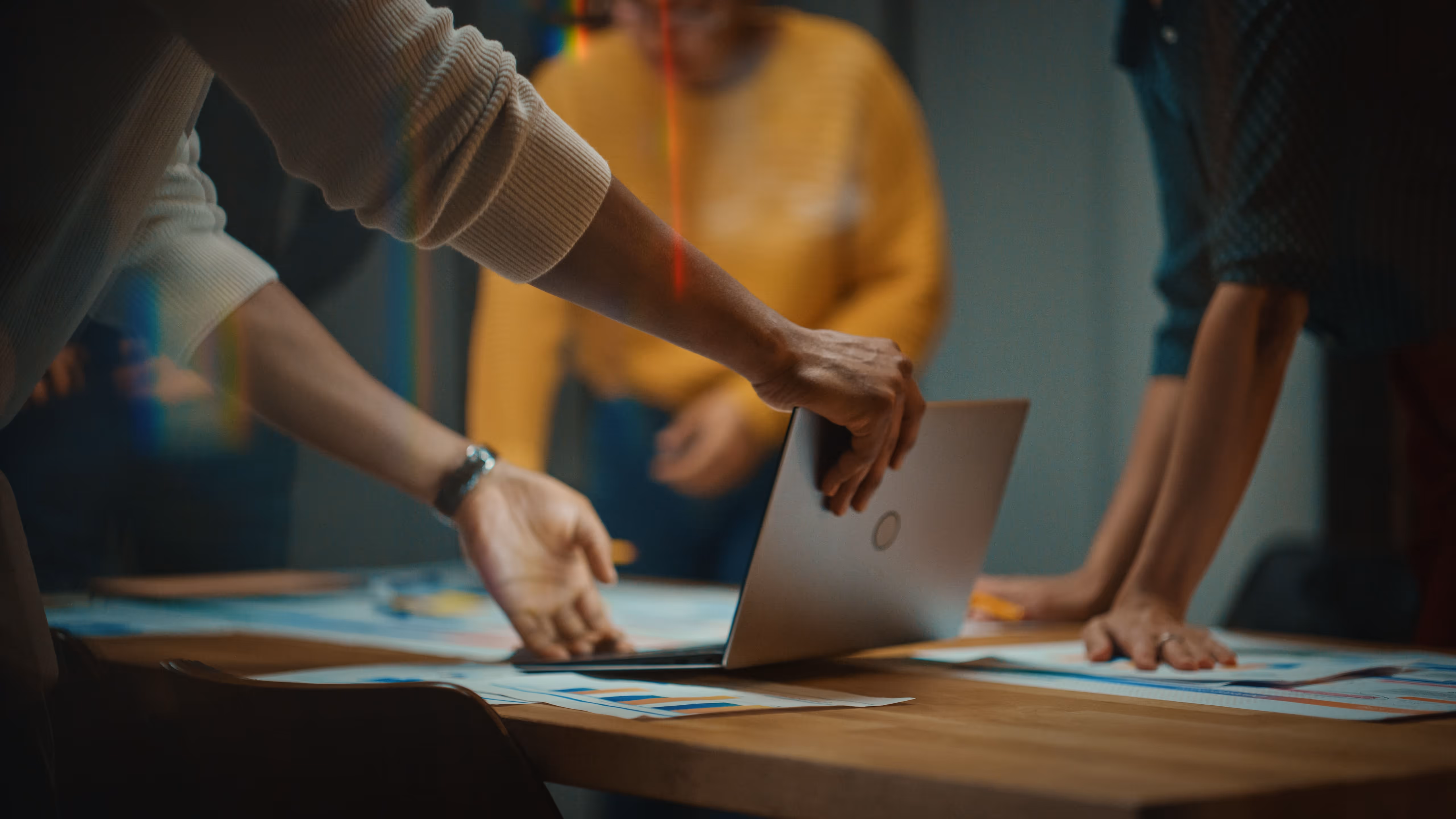People collaborating around a table with charts and opening a laptop.