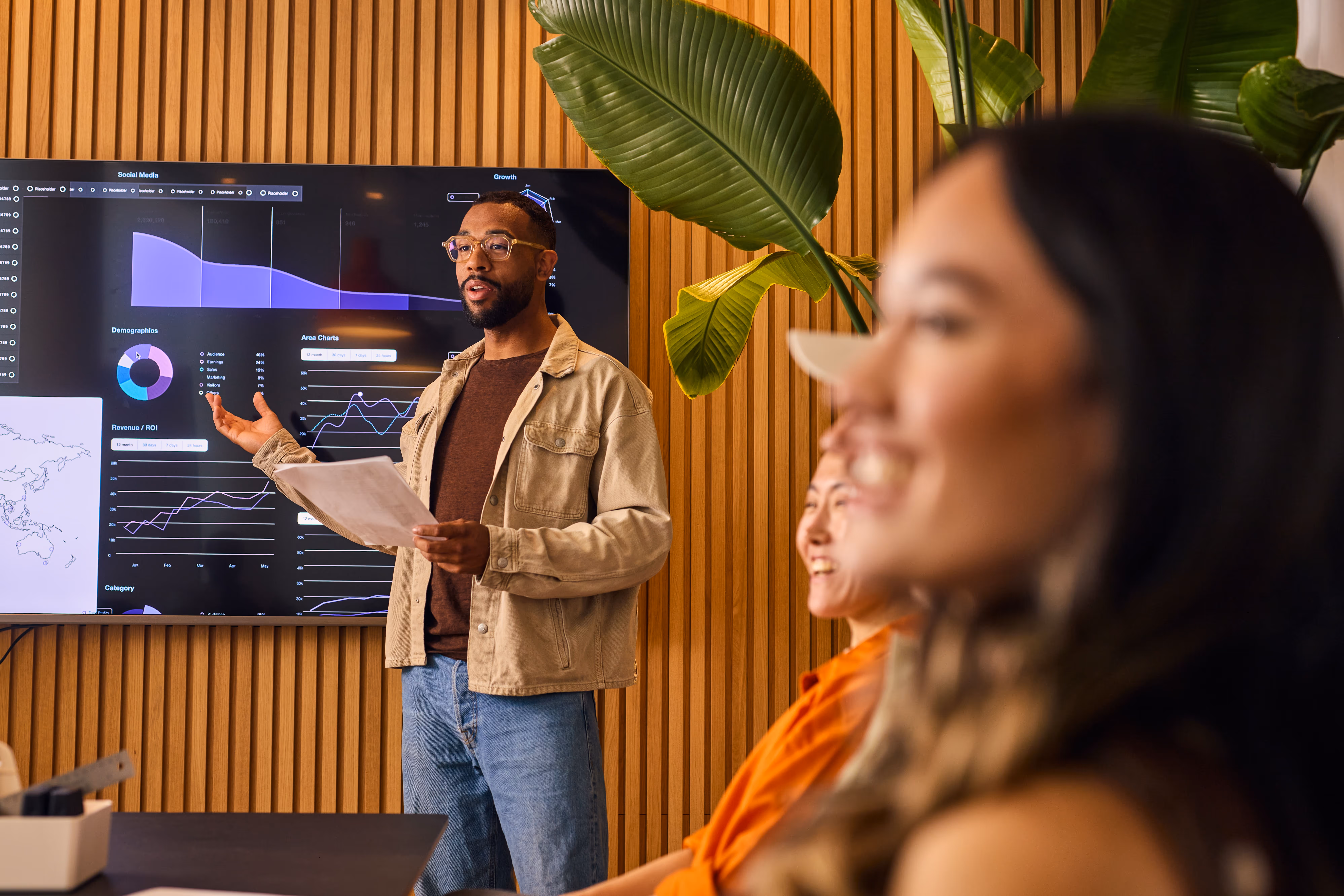 Man giving a presentation with charts on a screen while two colleagues listen and smile in a modern office.