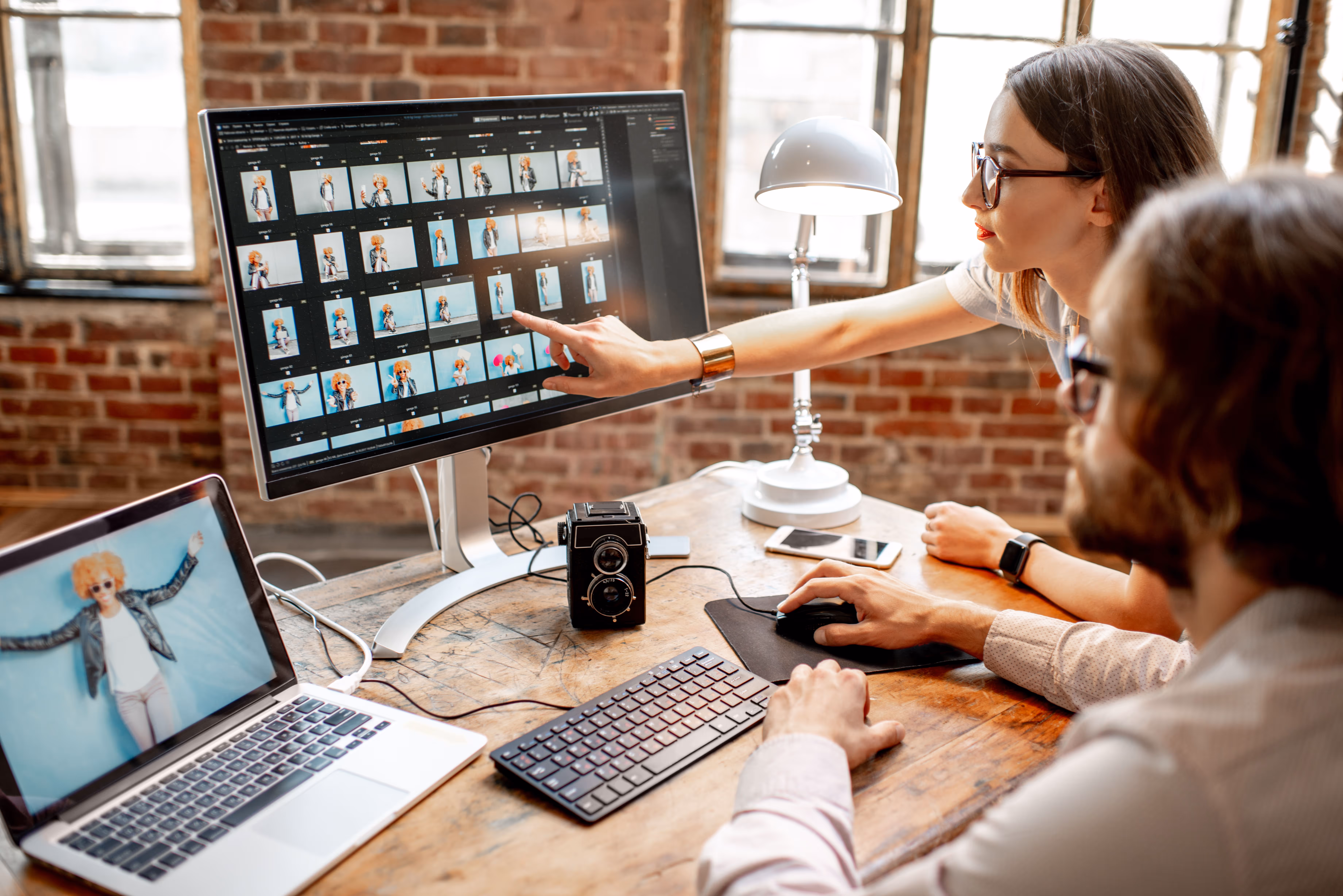 Two people editing and reviewing photo thumbnails on a large monitor in a rustic office, with a laptop displaying a photo and a vintage camera on the desk.