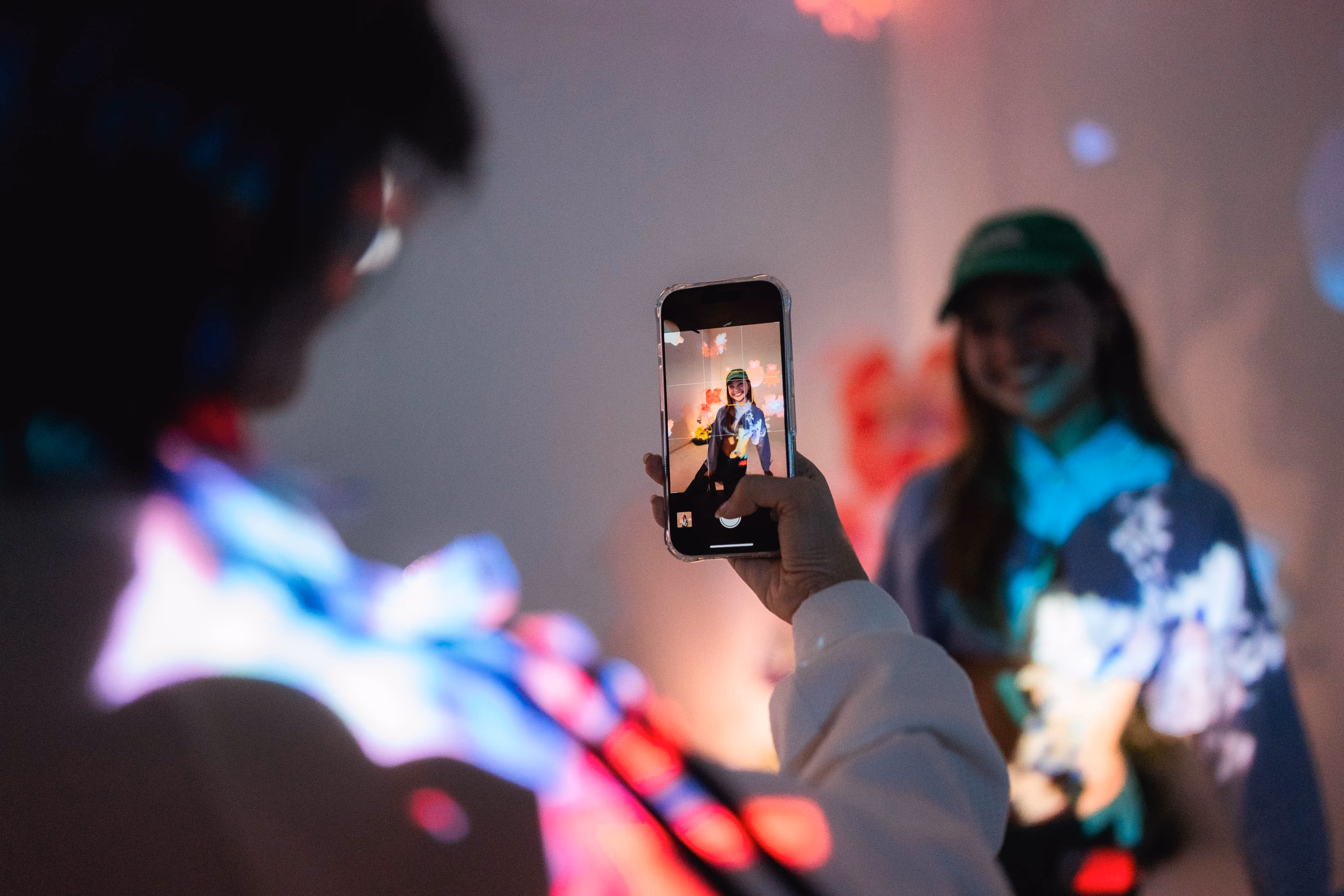 Person taking a photo with a smartphone of a smiling woman wearing a green cap near a wall with colorful light projections.