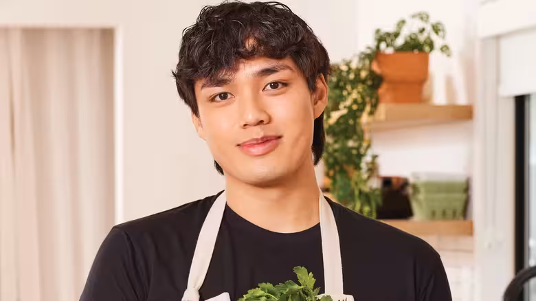 Young man with black hair wearing a black shirt and apron, smiling in a kitchen holding fresh greens.