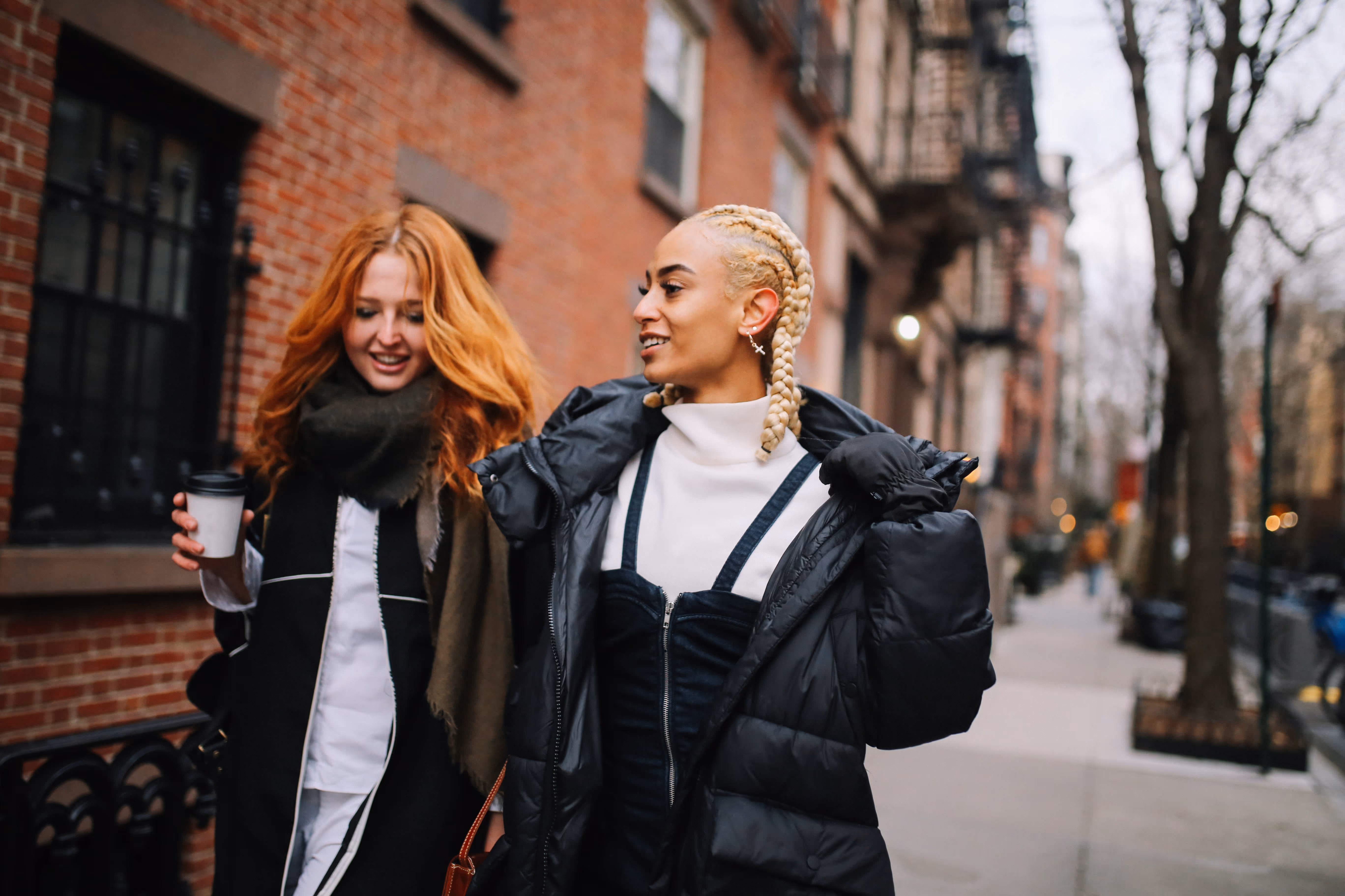 Two women walking and smiling on a city sidewalk, one holding a coffee cup and the other wearing a black puffer jacket with braided blonde hair.
