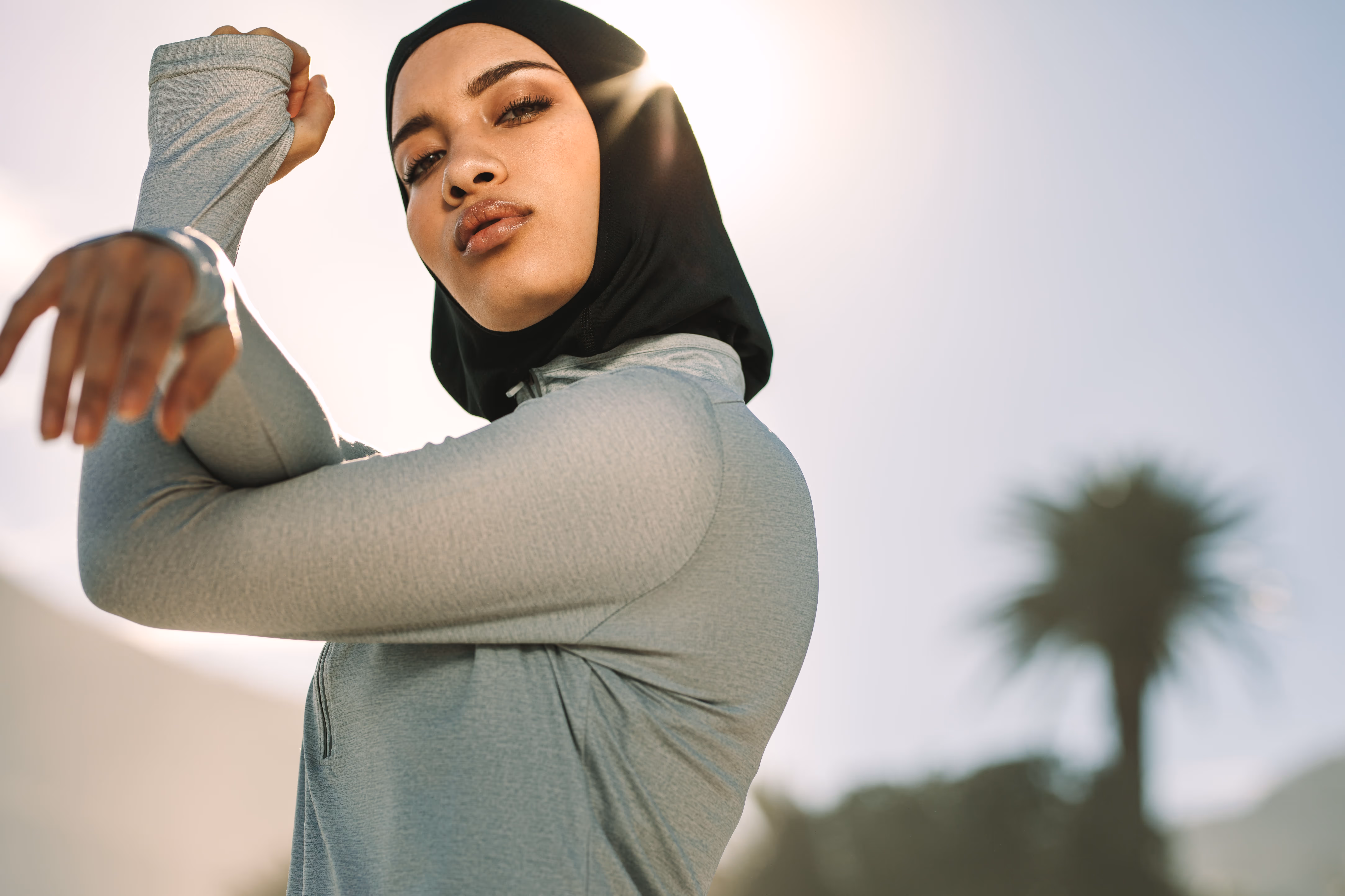 Confident woman in a black hijab and gray athletic wear stretching her arm outdoors with sunlight behind.