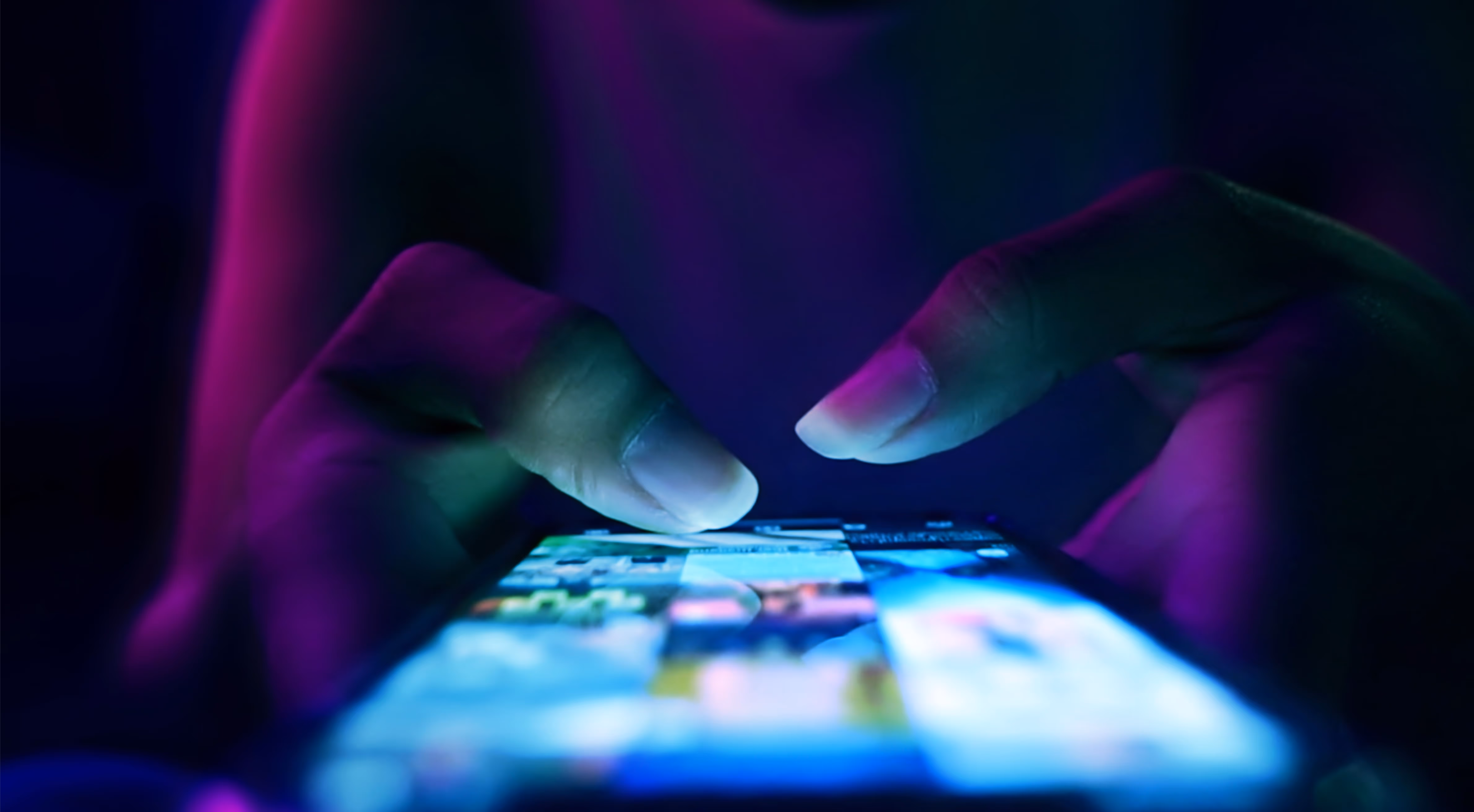 Close-up of hands using a smartphone with colorful screen in low light with blue and purple tones.
