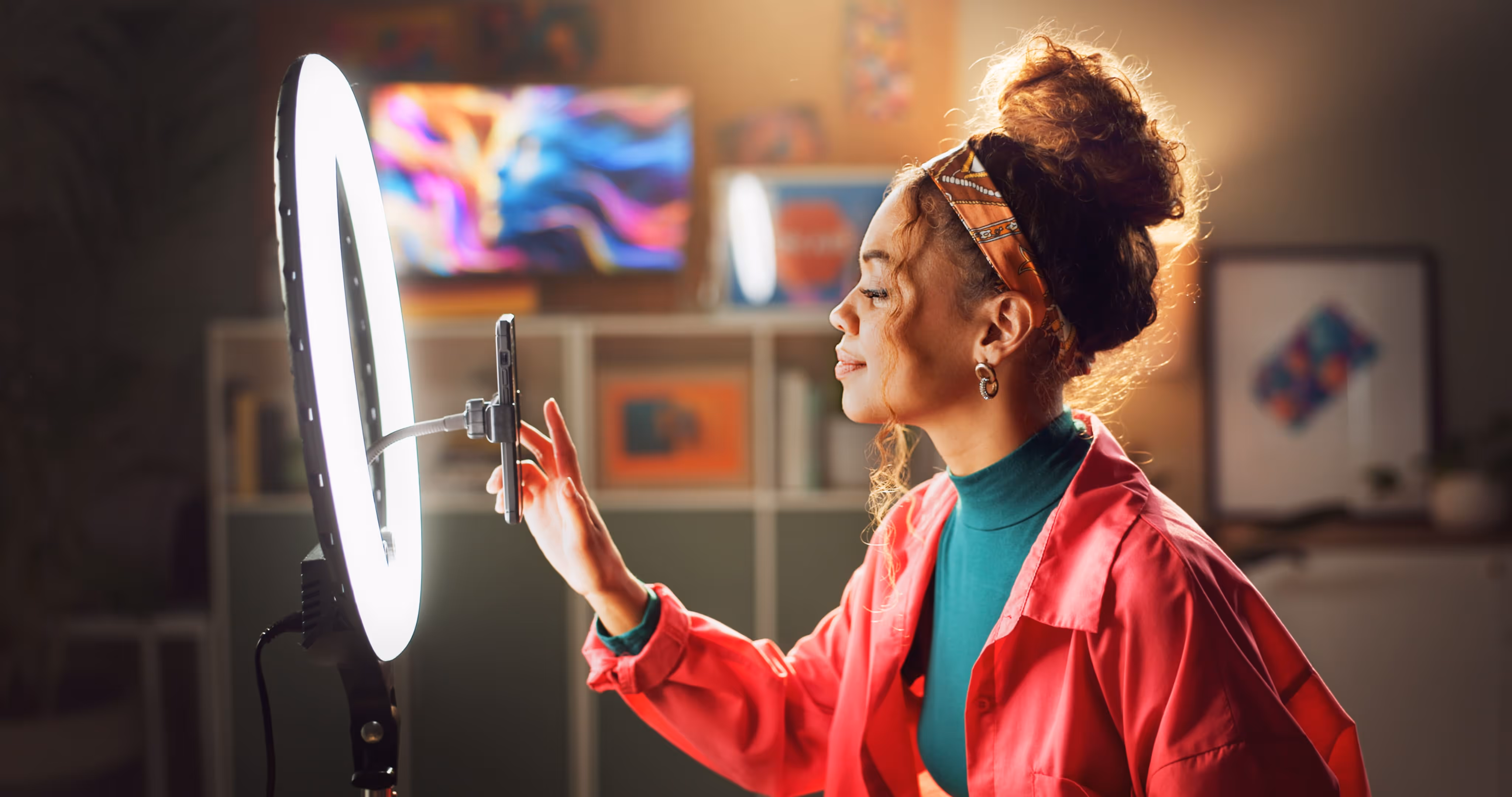 Young woman with curly hair tied up in a headband using a smartphone mounted on a ring light in a cozy room.