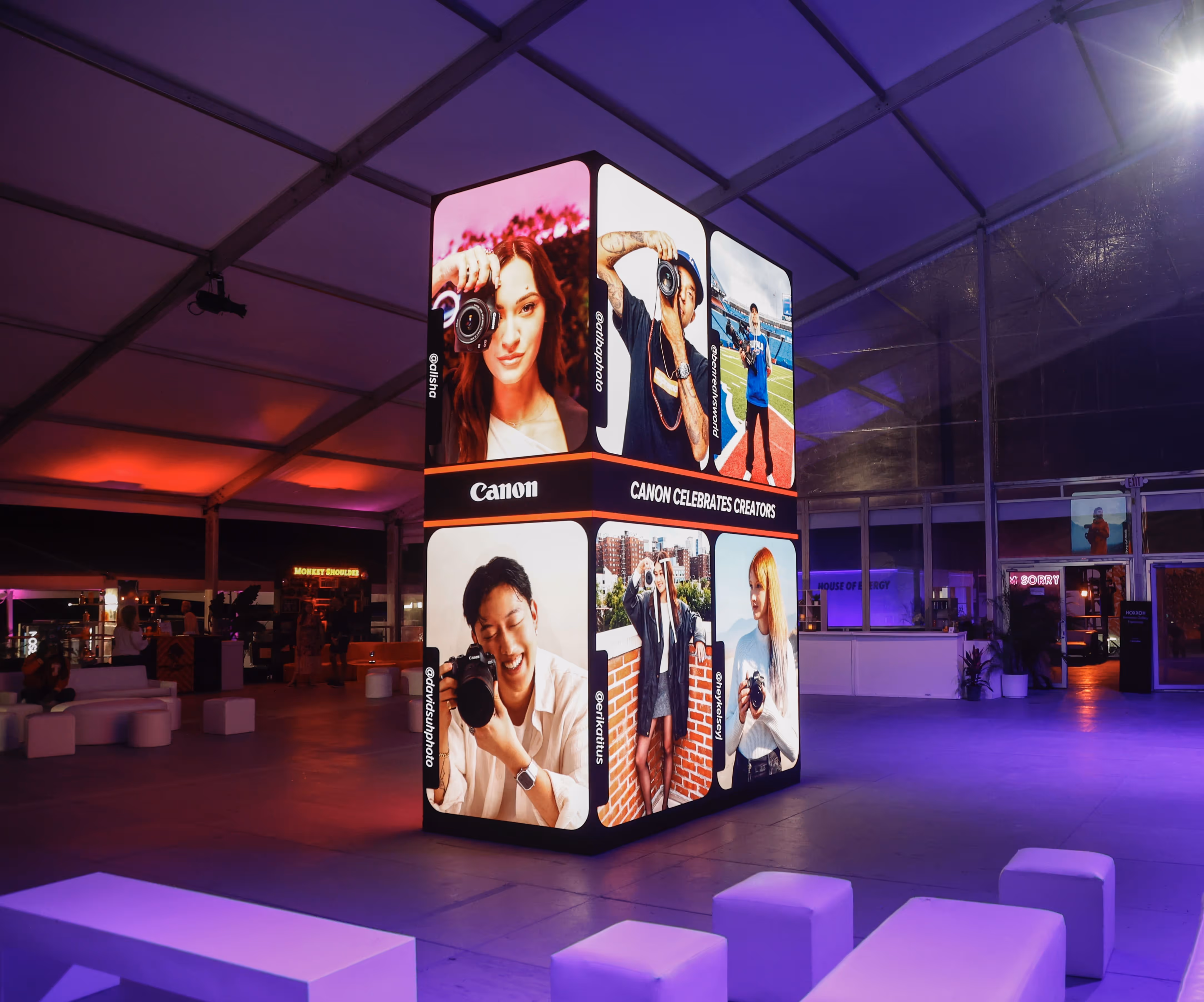 Illuminated cube-shaped display featuring portraits of six photographers, with Canon logo and 'Canon Celebrates Creators' text in a dimly lit indoor event space.
