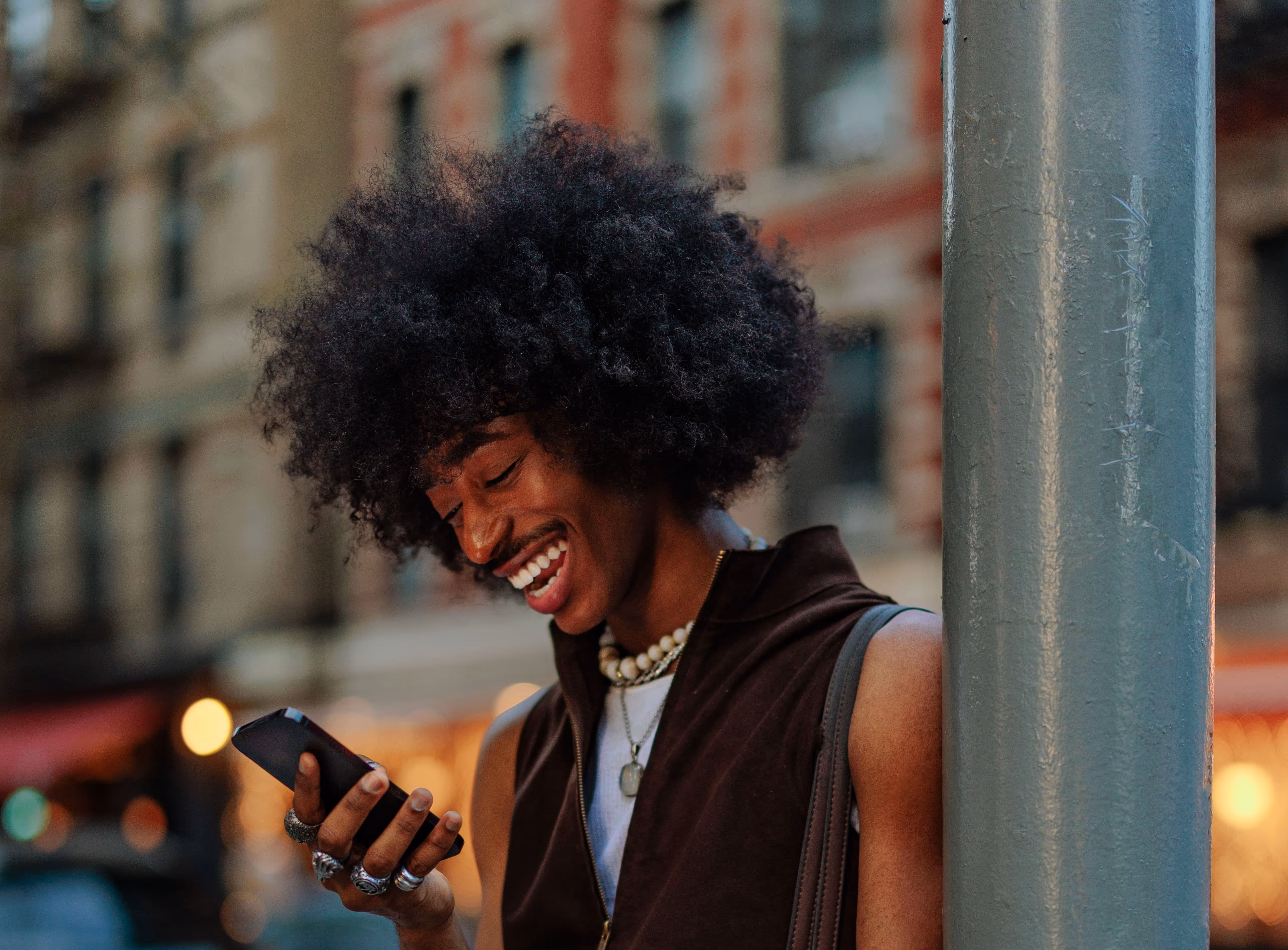 Smiling person with afro hairstyle looking at a smartphone on a city street.