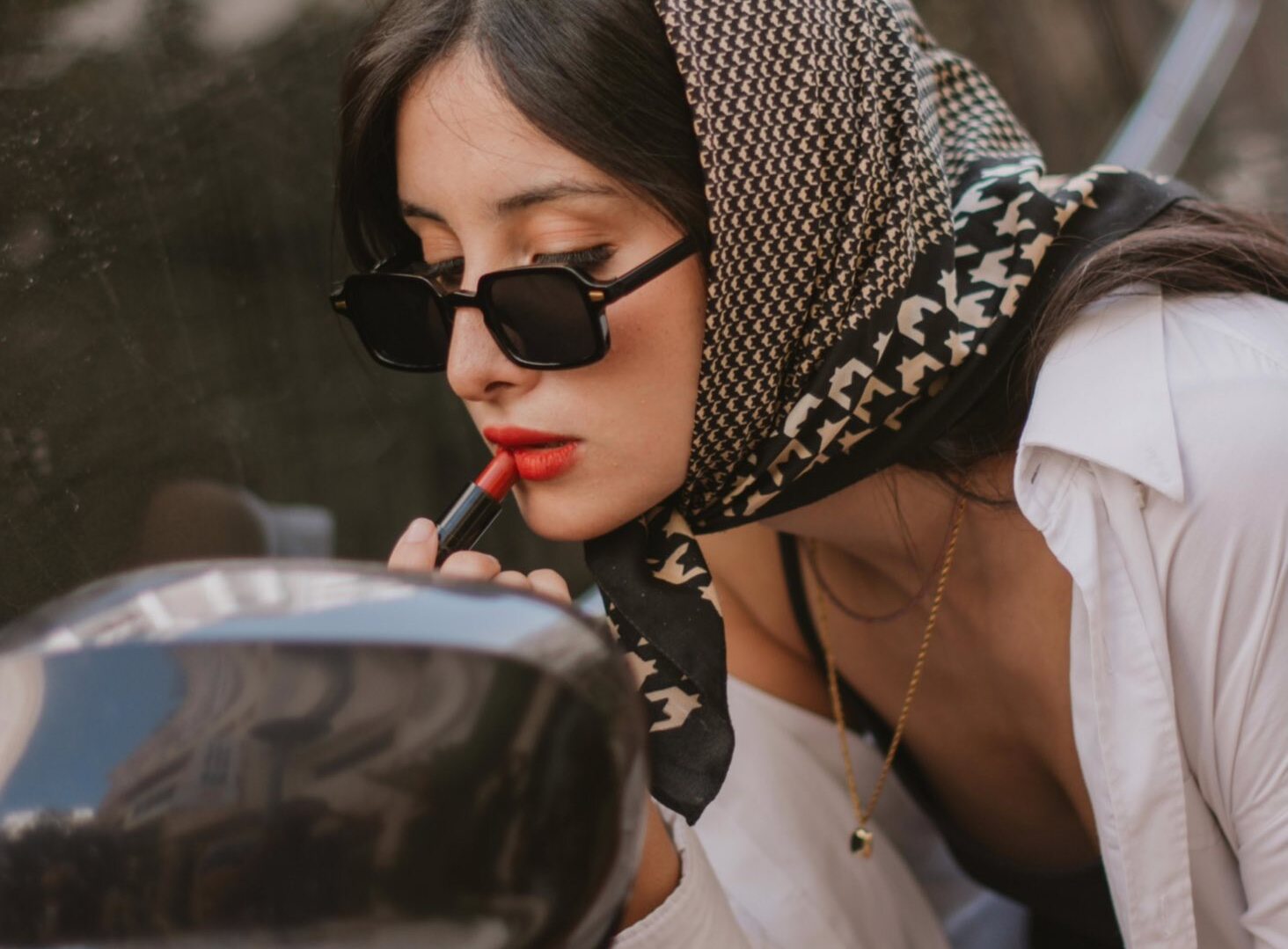 Woman wearing a patterned headscarf and sunglasses applying red lipstick while looking into a mirror.