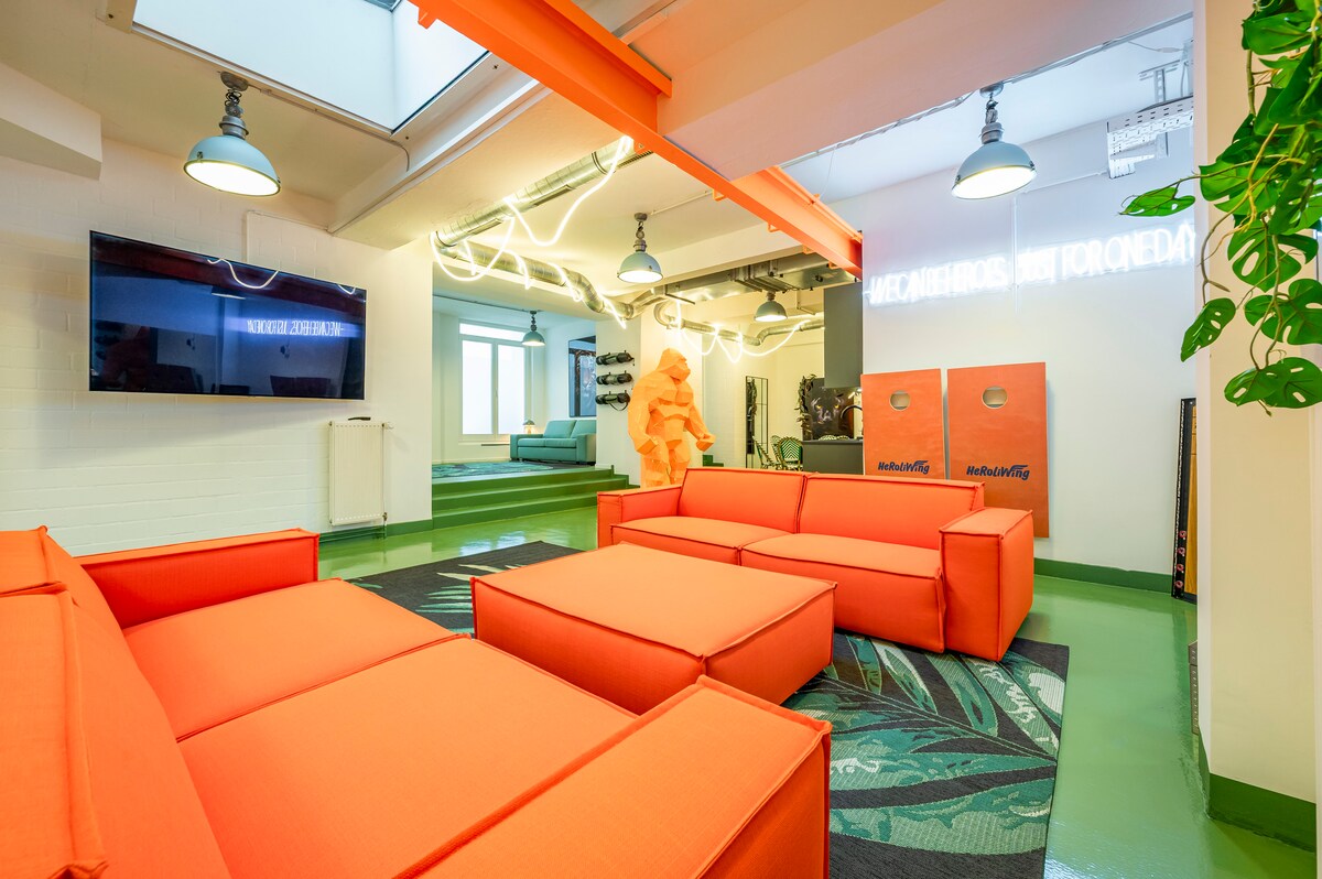 Moderner Loft-Wohnbereich mit knallorangenen Sofas und Hocker auf grünem Boden, Oberlicht und Industrial-Lampen, großer TV an der Wand, Neon-Schriftzug und Cornhole-Boards im Hintergrund.