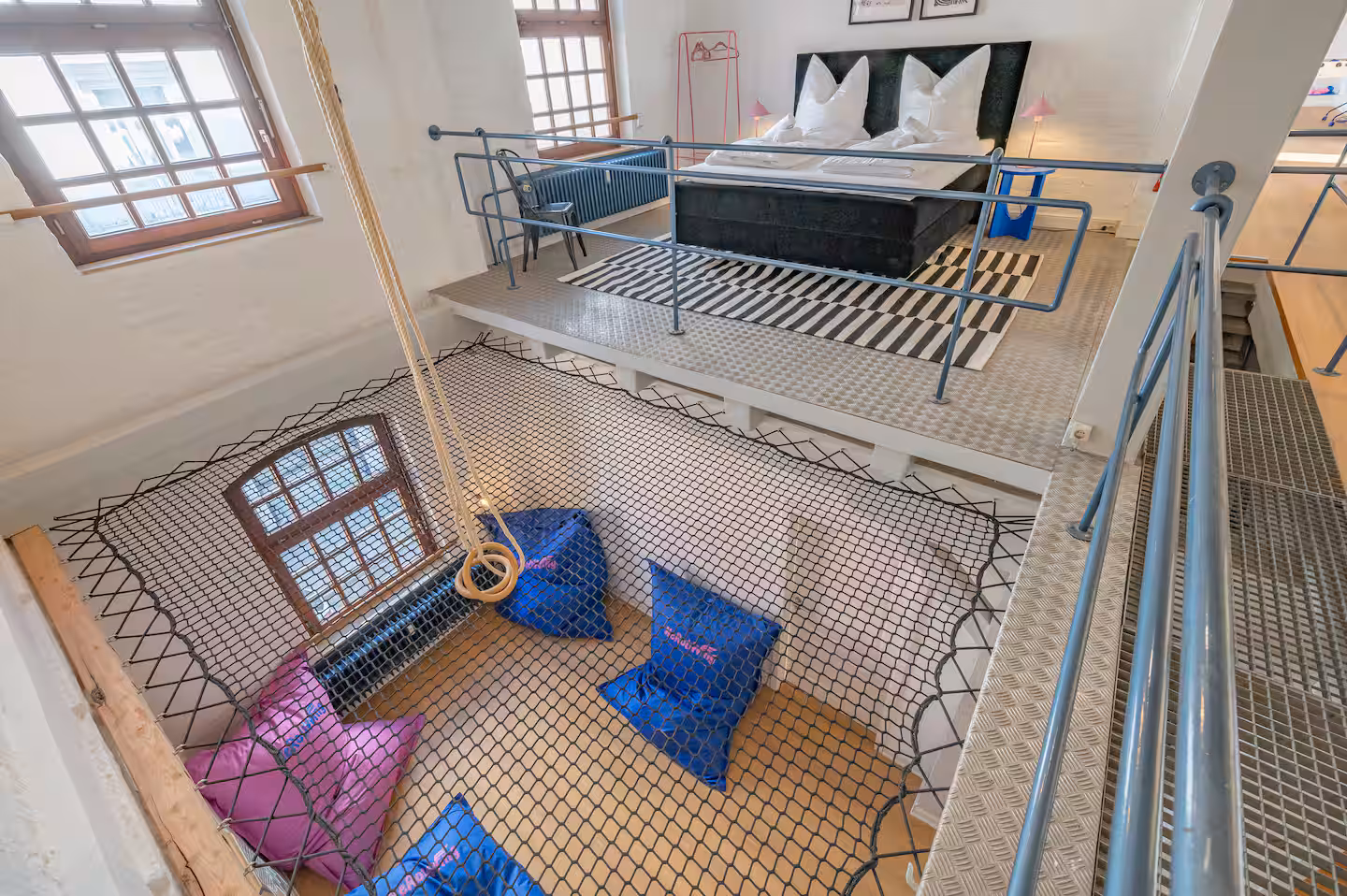 Helles Loft-Apartment mit Galerie-Schlafbereich: Doppelbett auf erhöhter Plattform, industrielle Metall-Stege und ein großes Hängemattennetz mit Sitzkissen unter hohen Fenstern.