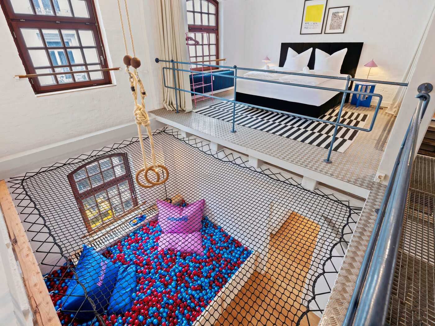 Helles Loft-Apartment mit Galerie-Schlafbereich: Doppelbett auf erhöhter Plattform, industrielle Metall-Stege und ein großes Hängemattennetz mit Sitzkissen unter hohen Fenstern.