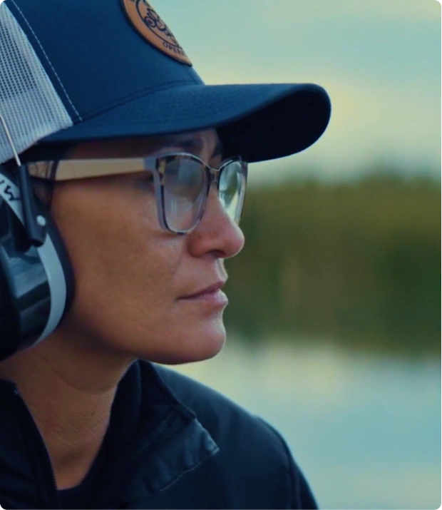 Side profile of a person wearing glasses, a dark cap, and large headphones against a blurred outdoor background.