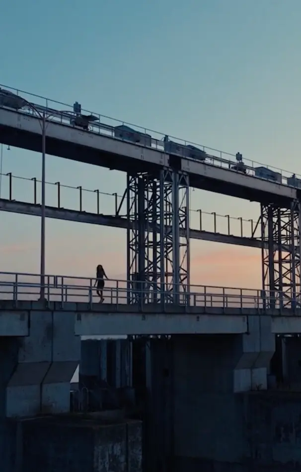 A person walking on a bridge or elevated walkway at sunset with vehicles parked on a higher level above them.