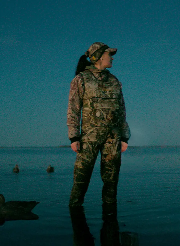 Nadina in camouflage gear standing in shallow water at dusk with ducks nearby.