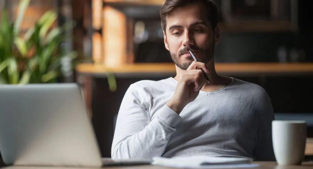 a man looking at a laptop, holding a pen thinking