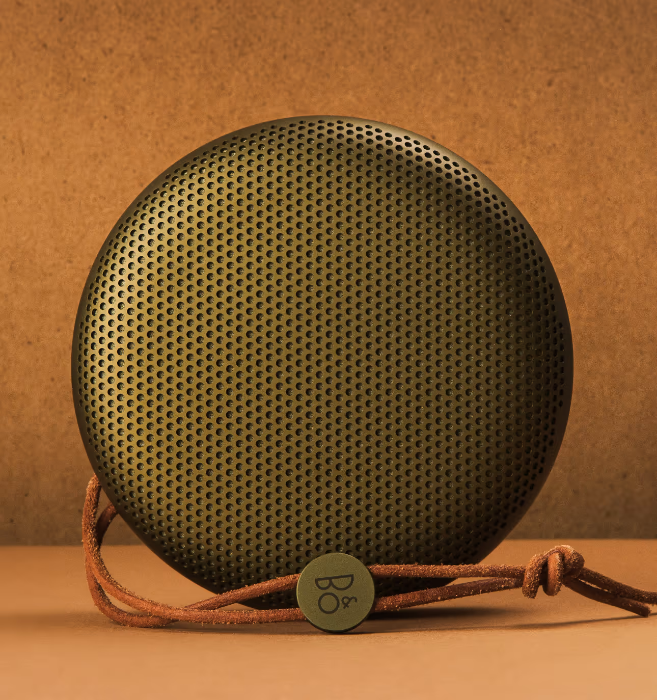 Green circular portable speaker with a leather strap and circular branded tag against a brown background.