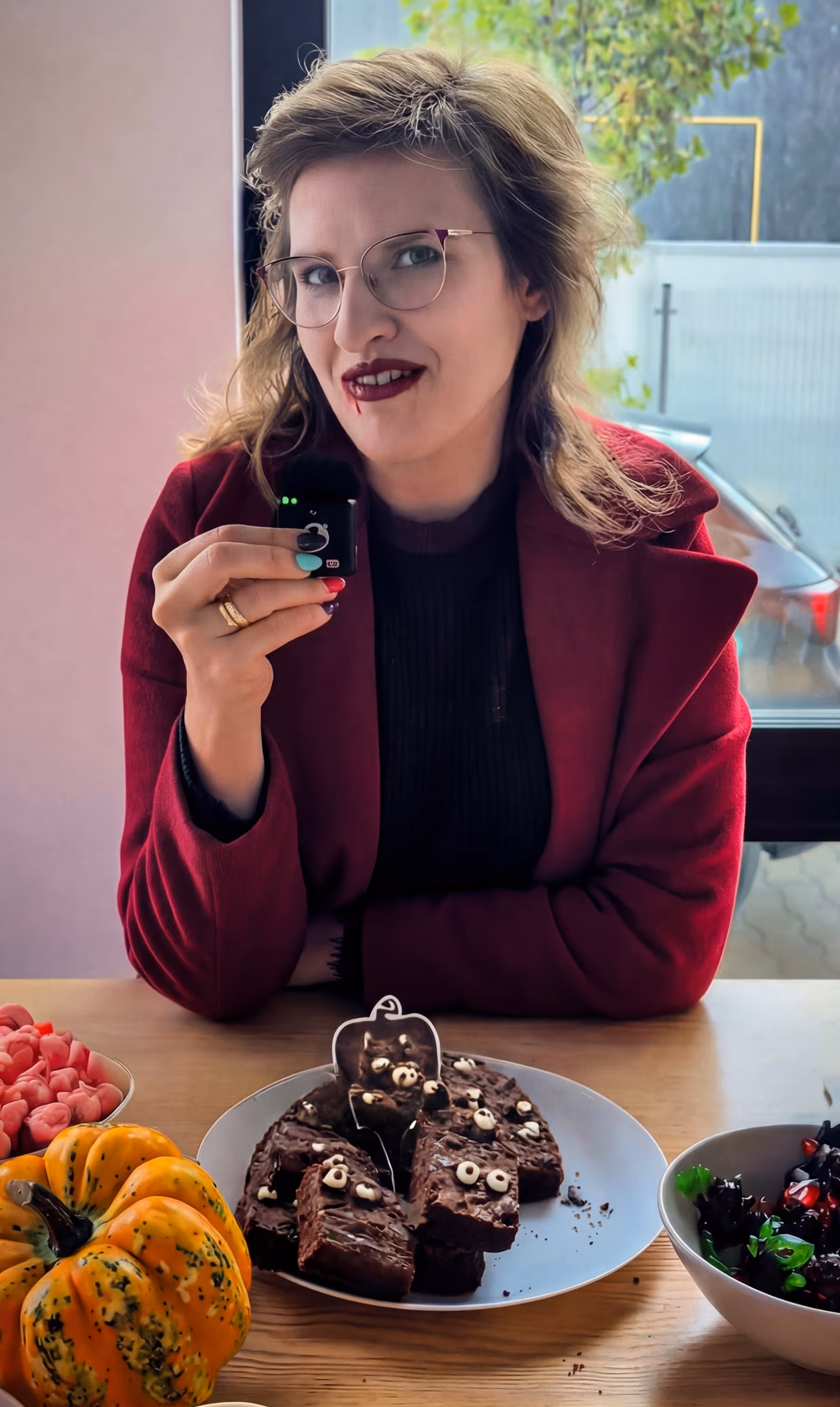 Kobieta w bordowym płaszczu z krwawymi ustami trzyma mikrofon, przed nią na stole talerz z ciastkami na Halloween z oczami i misterną dekoracją, obok dynia i miska z sałatką.