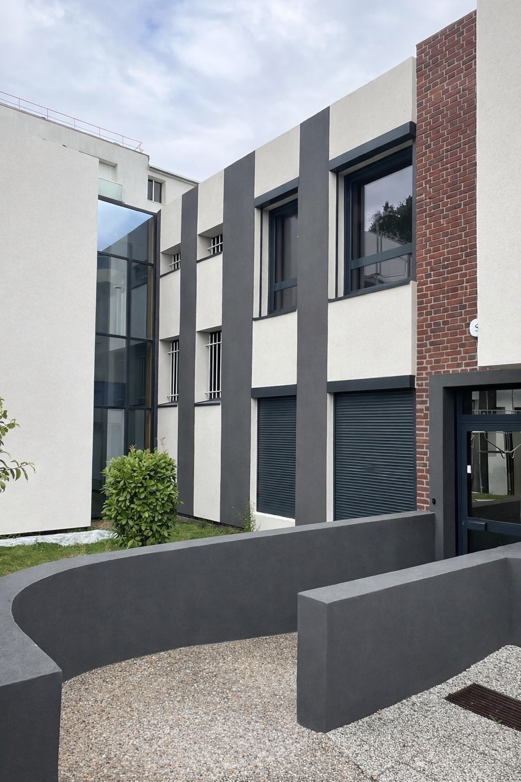 Façade moderne du bâtiment avec des rayures verticales grises et blanches, des fenêtres à volets, une section d'angle vitrée et une rampe incurvée en béton gris.