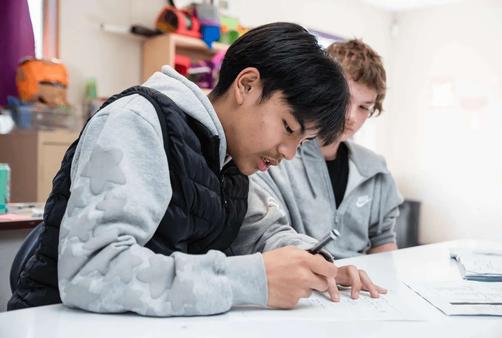 Two students in hoodies focused on writing and studying at a white table in a classroom.