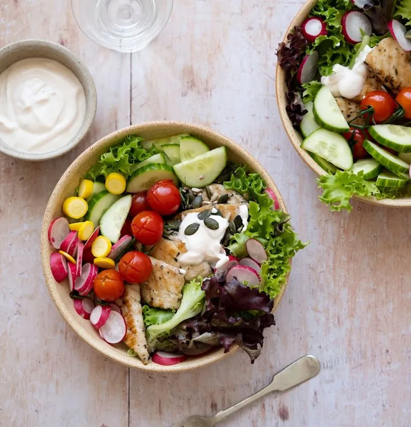 Two bowls of fresh salad with grilled chicken, cherry tomatoes, cucumber slices, radish, corn, mixed greens, and a dollop of creamy dressing topped with pumpkin seeds, accompanied by a small bowl of white sauce.