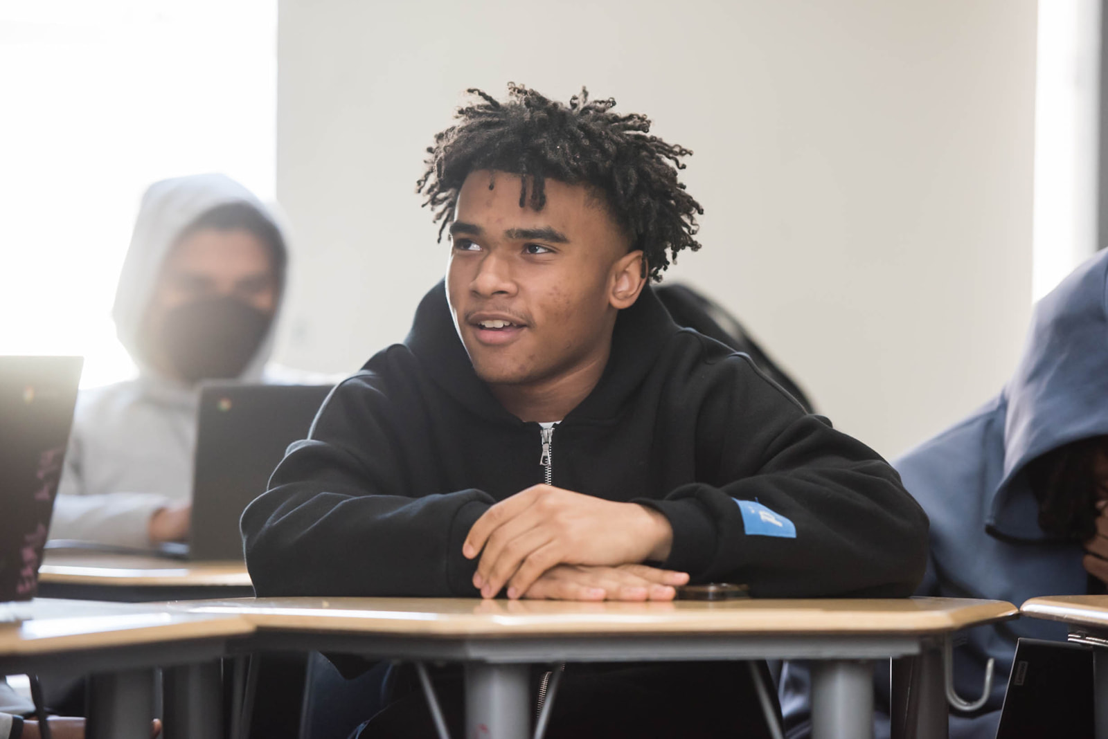 A teenage boy with short twisted hair sits at a classroom desk wearing a black hoodie. He rests his arms on the desk and looks off to the side with an attentive expression, while another student sits blurred in the background.