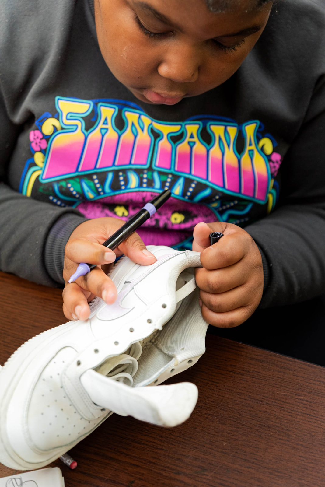 Child wearing a colorful Santana sweatshirt customizing a white Nike sneaker with a purple marker.