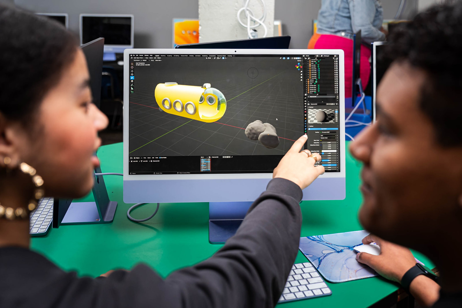 Two people collaborate on a 3D modeling project displayed on a computer screen showing a yellow submarine and rocks.