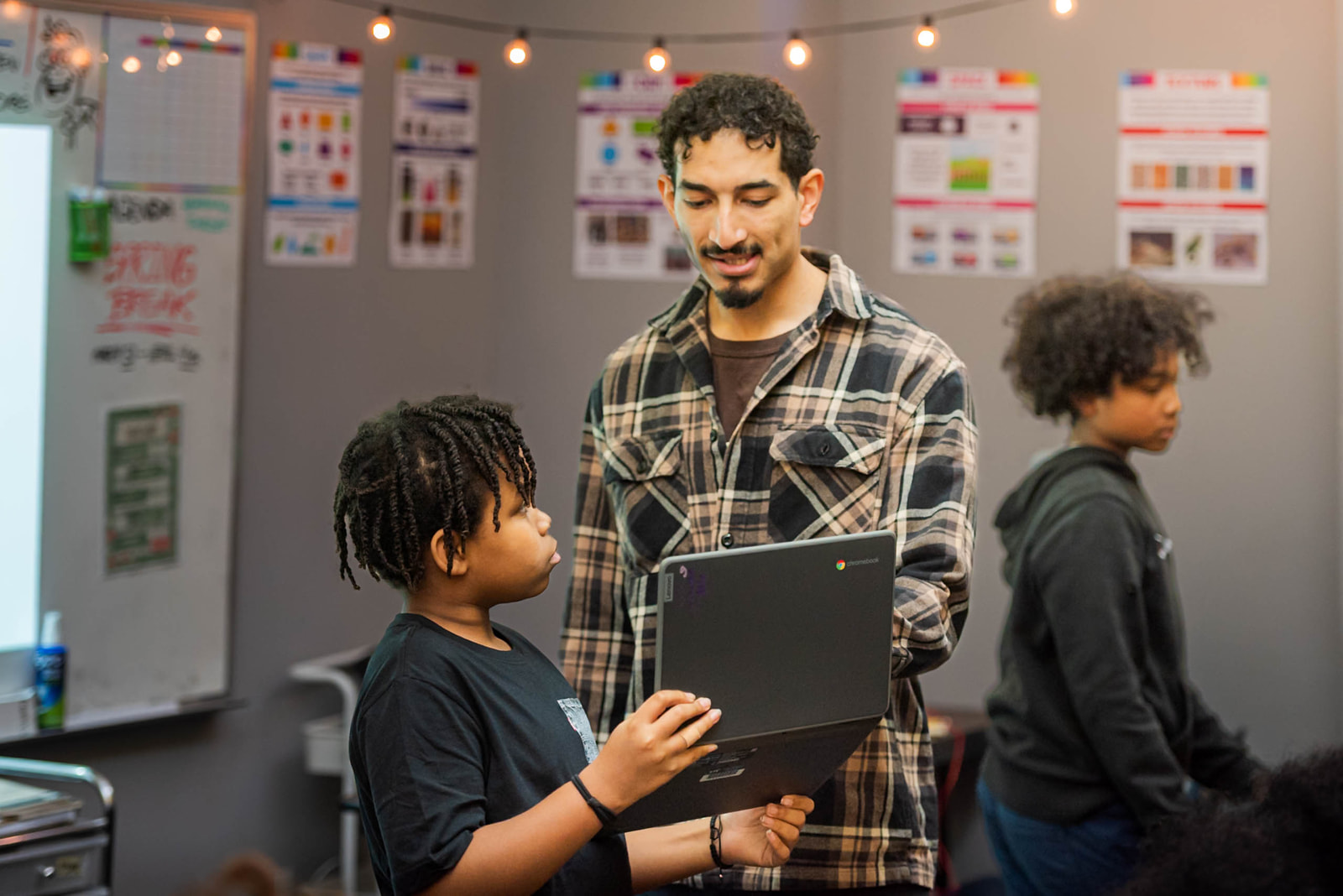 Teacher in a plaid shirt helping a student holding a Chromebook in a classroom with another student nearby.
