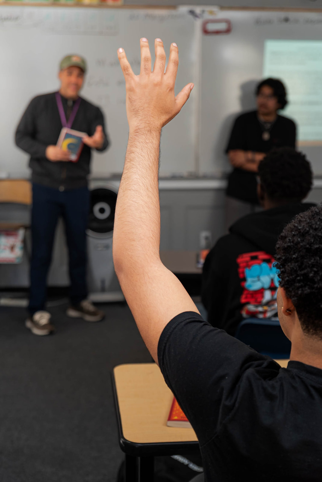 Student raising hand in classroom with teacher and another student in the background near a whiteboard.