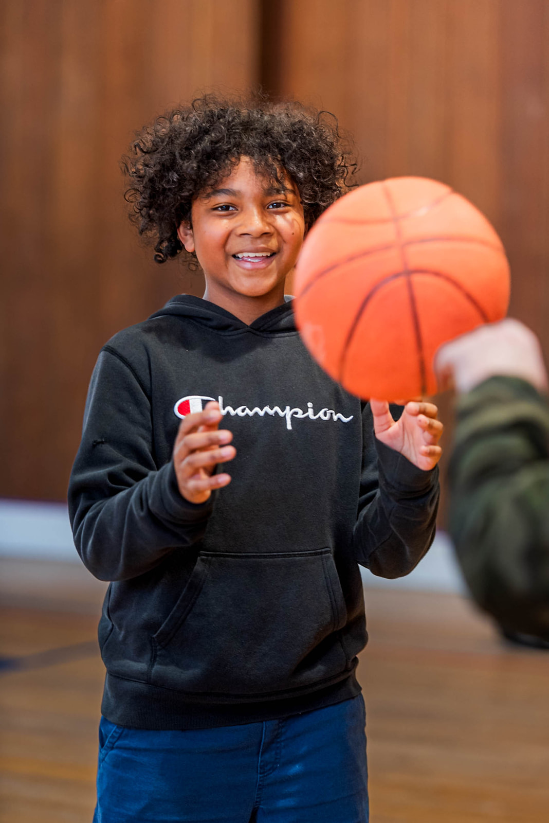 Smiling child in a black Champion hoodie catching an orange basketball indoors.