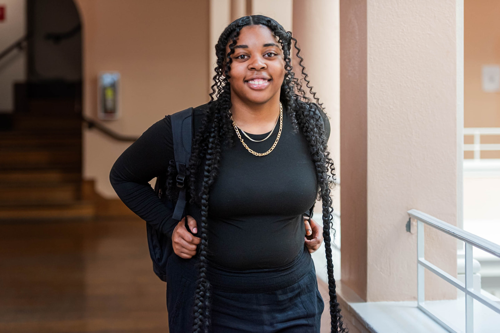 Smiling young woman with long curly braids wearing black clothes and a backpack indoors.