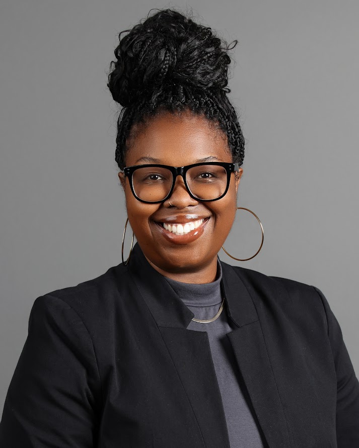 Smiling woman with braided hair in a bun, wearing large round earrings, black glasses, and a black blazer over a gray top against a gray background.
