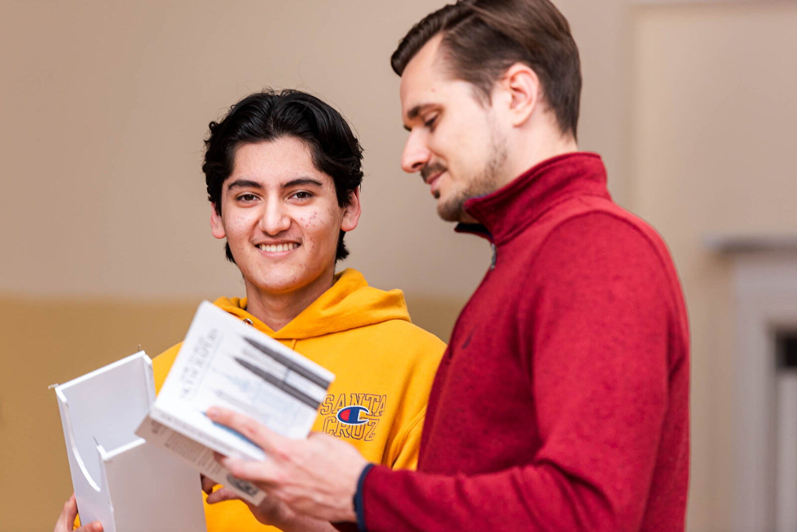 Two men reviewing documents or books together, one smiling at the camera wearing a yellow hoodie with Santa Cruz lettering, the other in a red sweater looking at the materials.