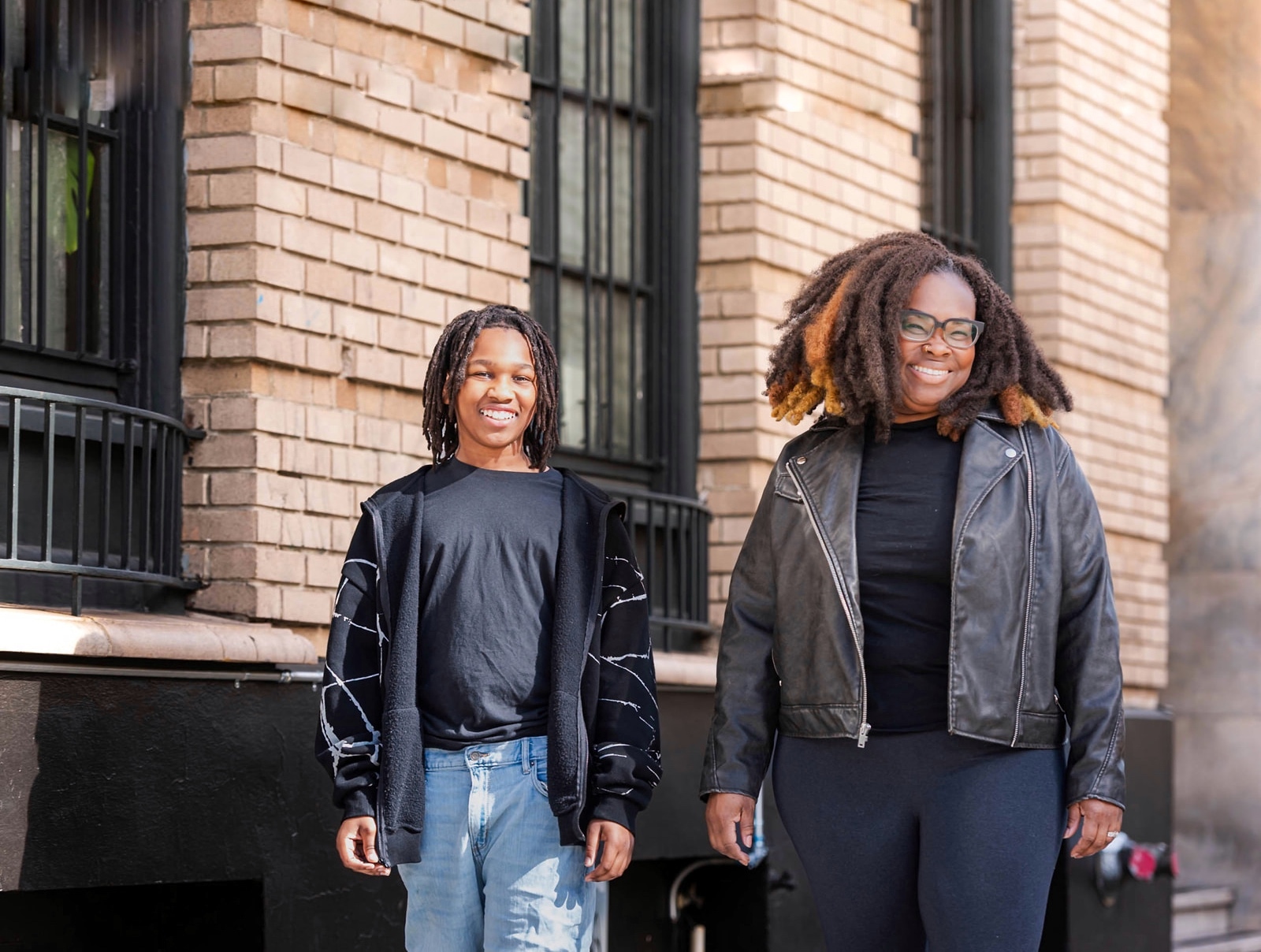 Two smiling people standing outside in front of a brick building, one wearing a black leather jacket and glasses, the other in a black and white hoodie and light blue jeans.