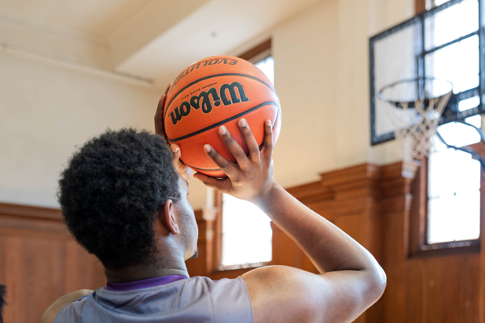 Back view of a basketball player holding a Wilson basketball preparing to shoot inside a gym with wooden wall panels and large windows.
