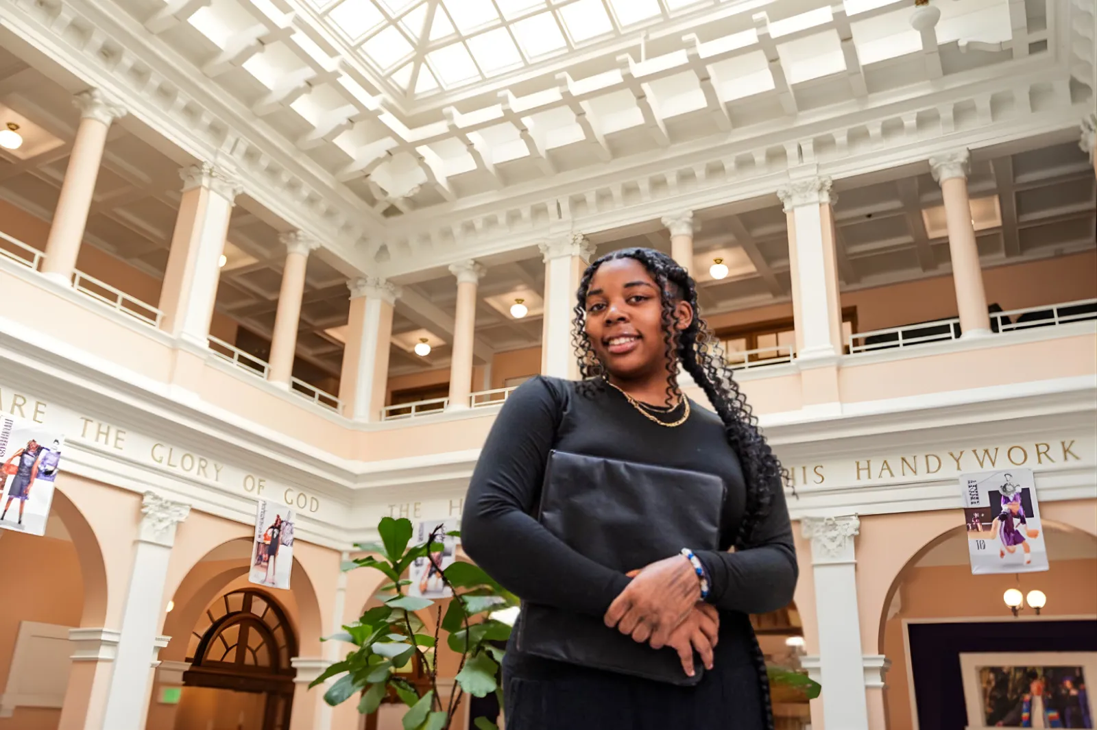 Smiling young woman with braided hair holding a folder inside a building with tall white columns and a skylight ceiling.