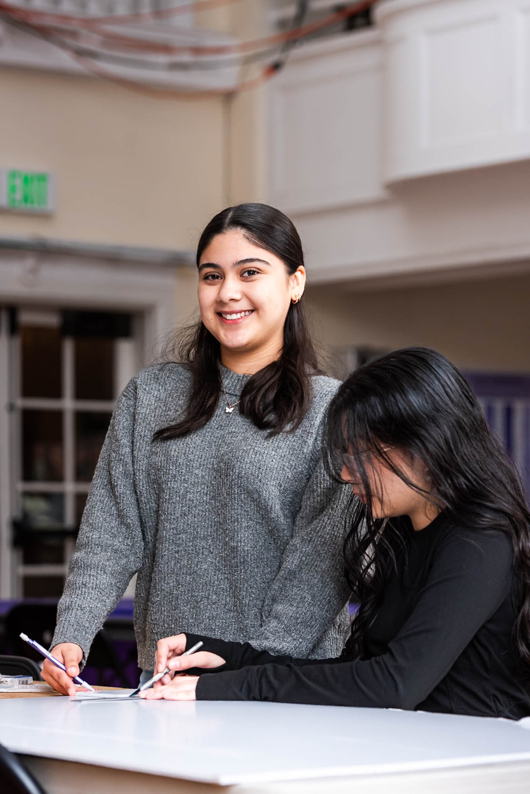 Two students smiling and pointing at a computer screen displaying a 3D yellow submarine model in a classroom.